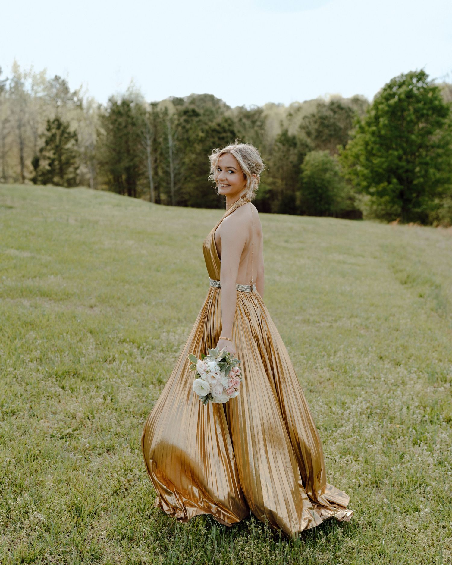 Someone in an elegant gold pleated evening gown stands in a grassy meadow holding a white floral bouquet.
