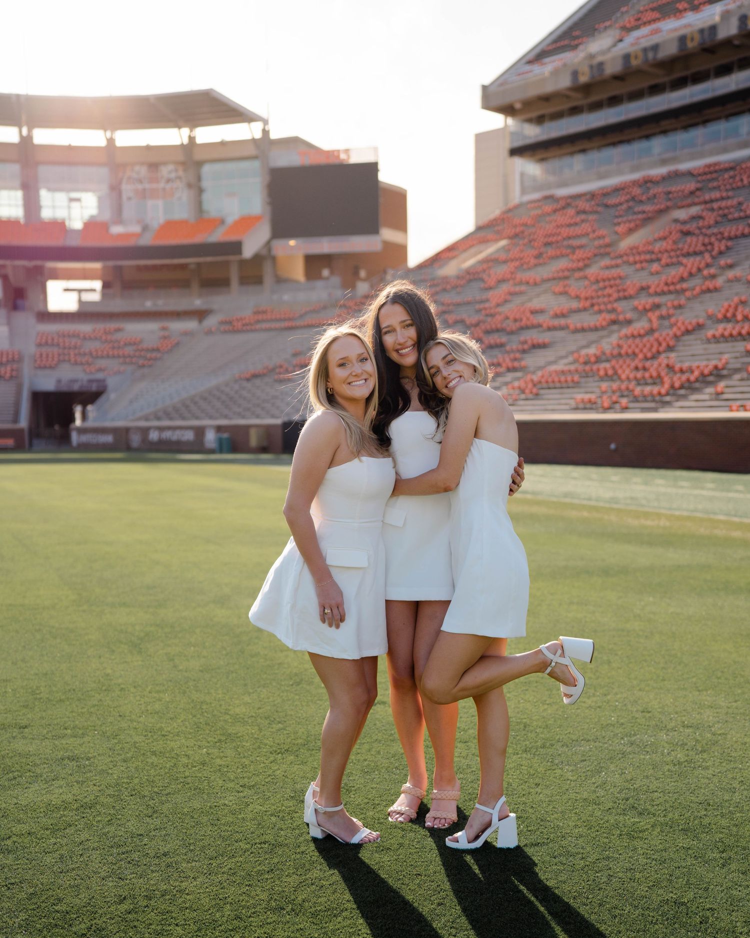 A group in matching white dresses pose together on a football field at sunset with stadium seating in background.