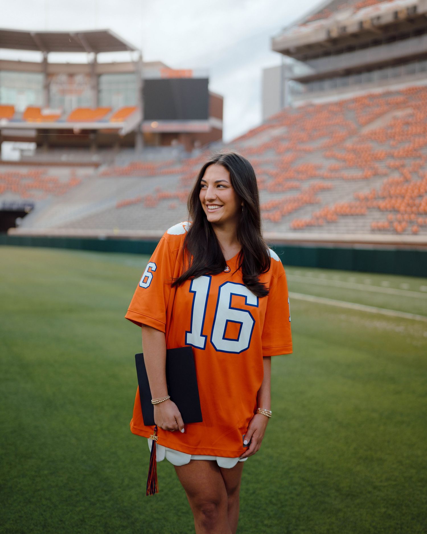 A graduate in an orange #16 jersey poses for a photo on a football field with stadium seating visible in the background.