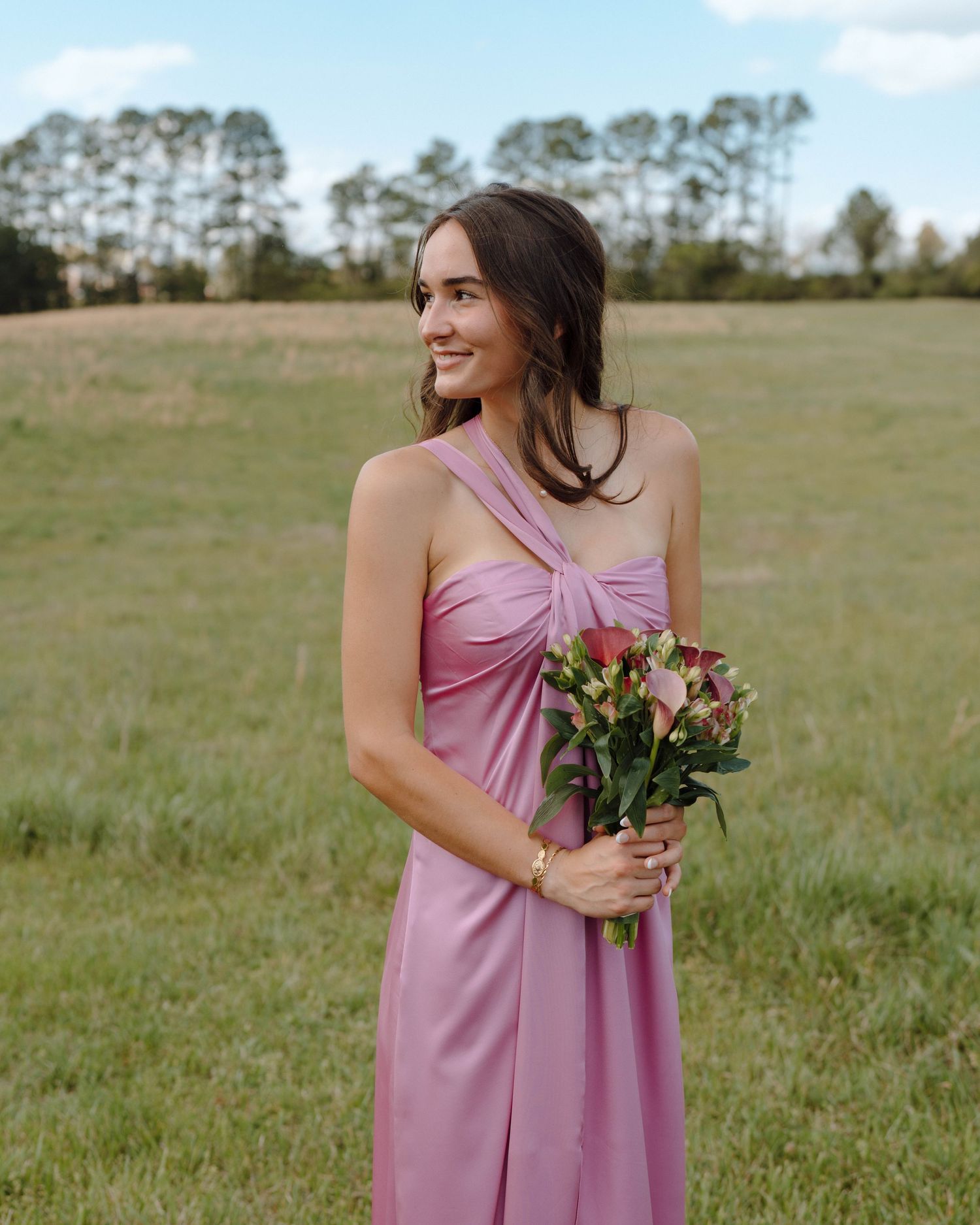 Woman in a pink bridesmaid dress holding a small bouquet of flowers smiles while standing in a grassy field with trees.