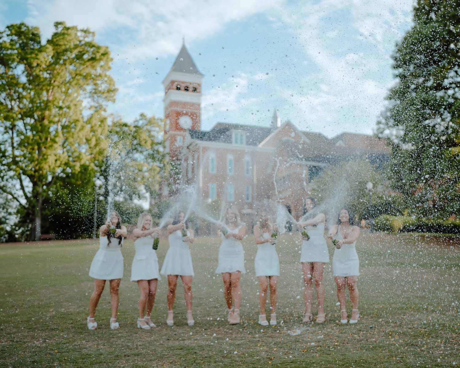 A group in white dresses celebrates on a lawn in front of a historic brick building with a bell tower.