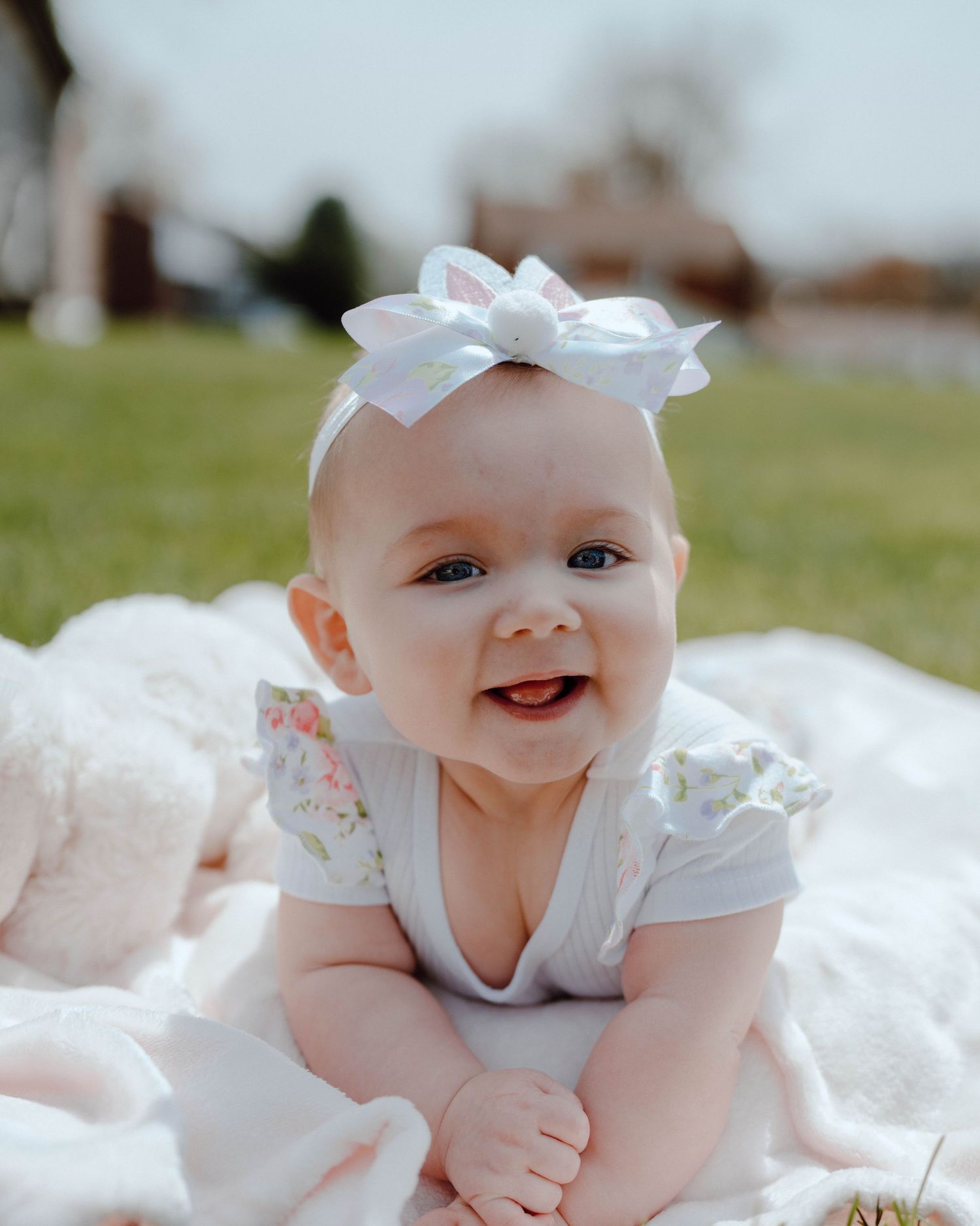 Baby wearing white outfit and bow smiles while laying on blanket outdoors on sunny day.
