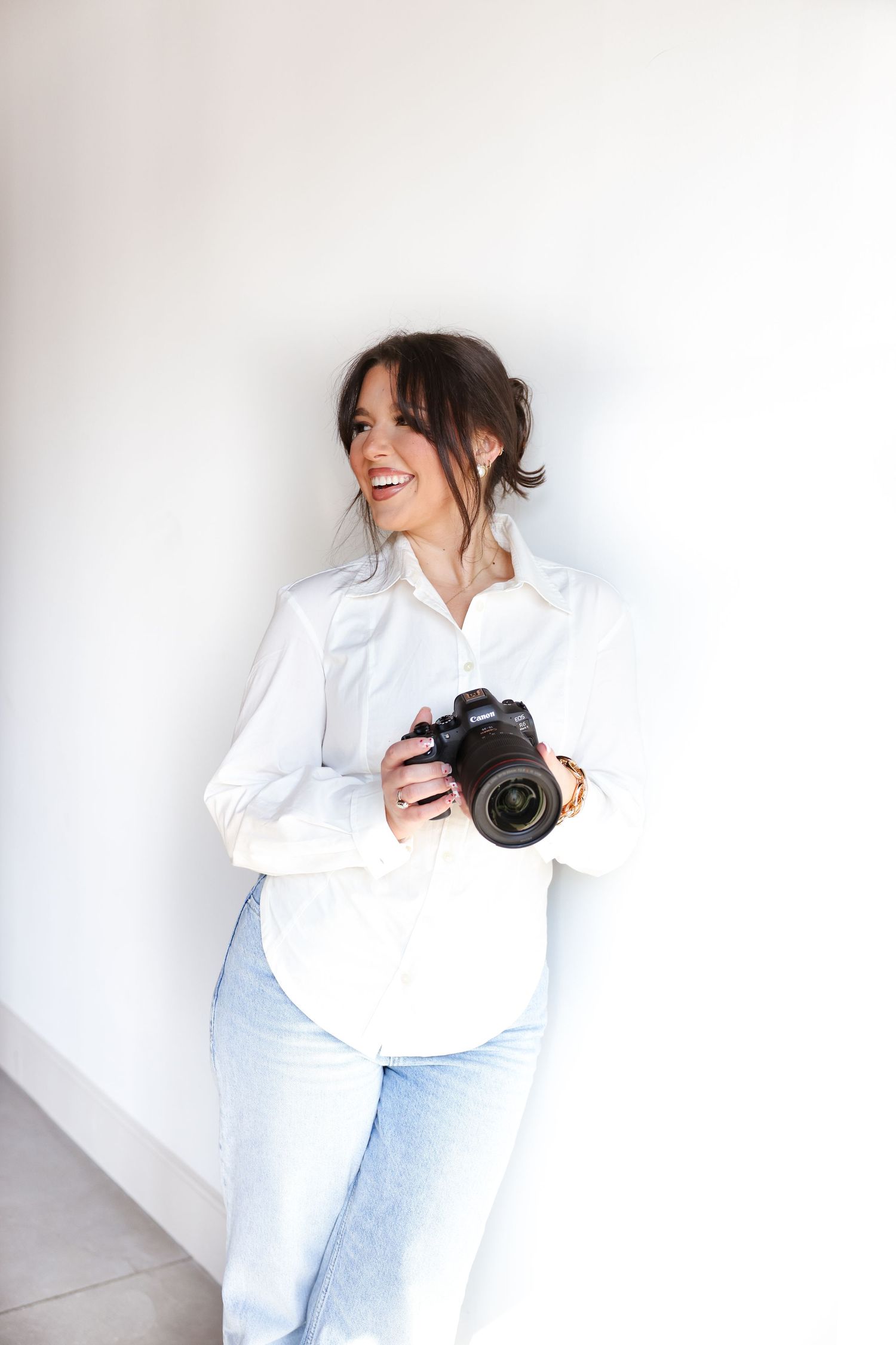 Professional photographer in white button-down shirt and light jeans holds digital camera while standing against white wall.