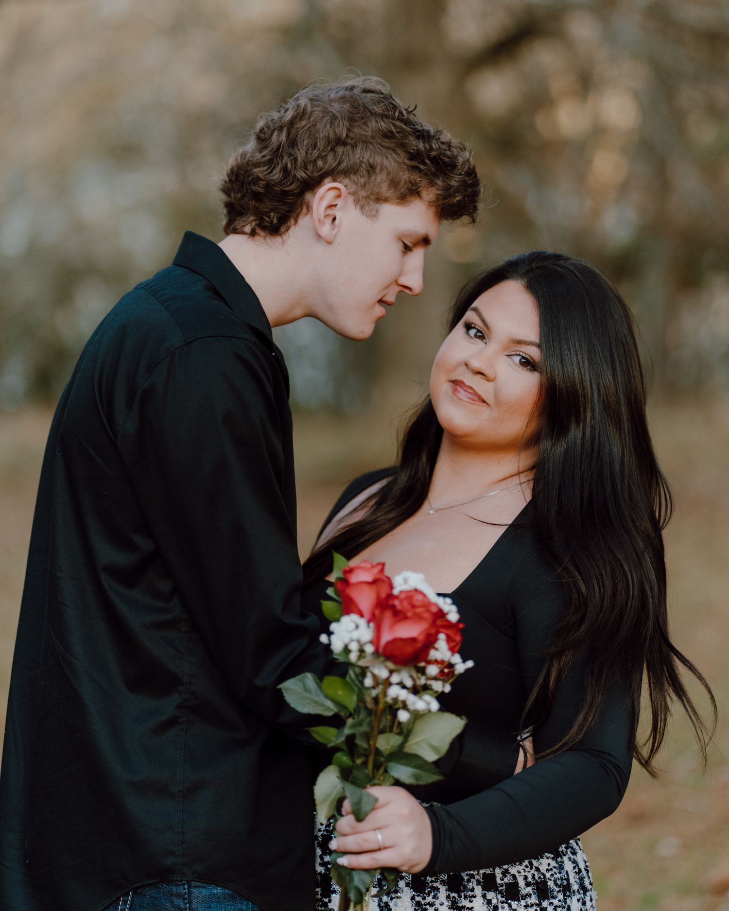 A romantic outdoor couple's portrait as they gaze at each other lovingly while holding red roses and baby's breath.