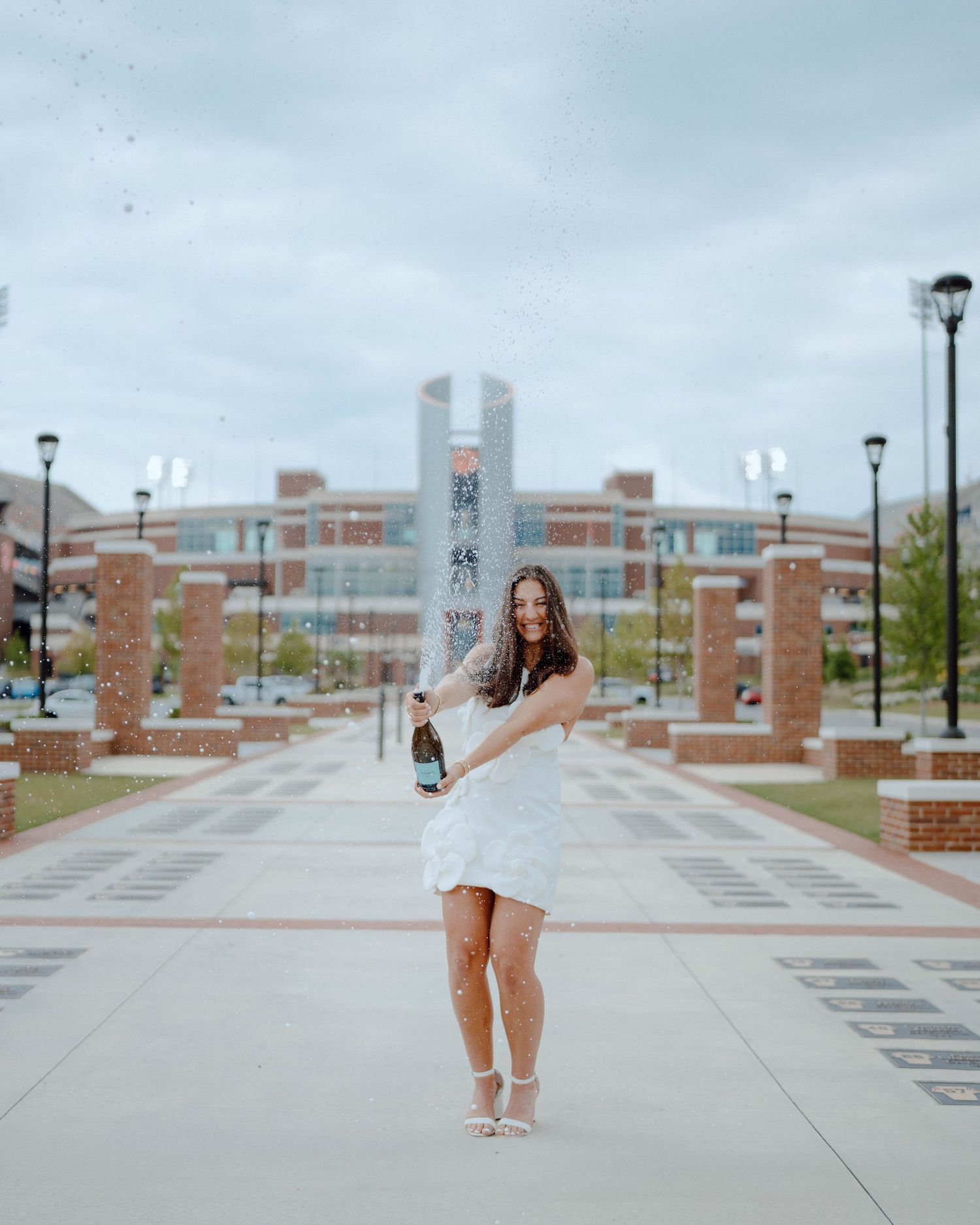 Graduate celebrating in white dress with champagne bottle on campus walkway under cloudy sky.
