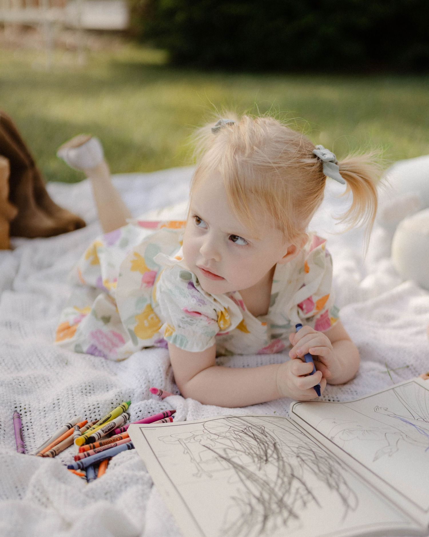 A small child lies on a white blanket outdoors with crayons and a coloring book on a sunny day.