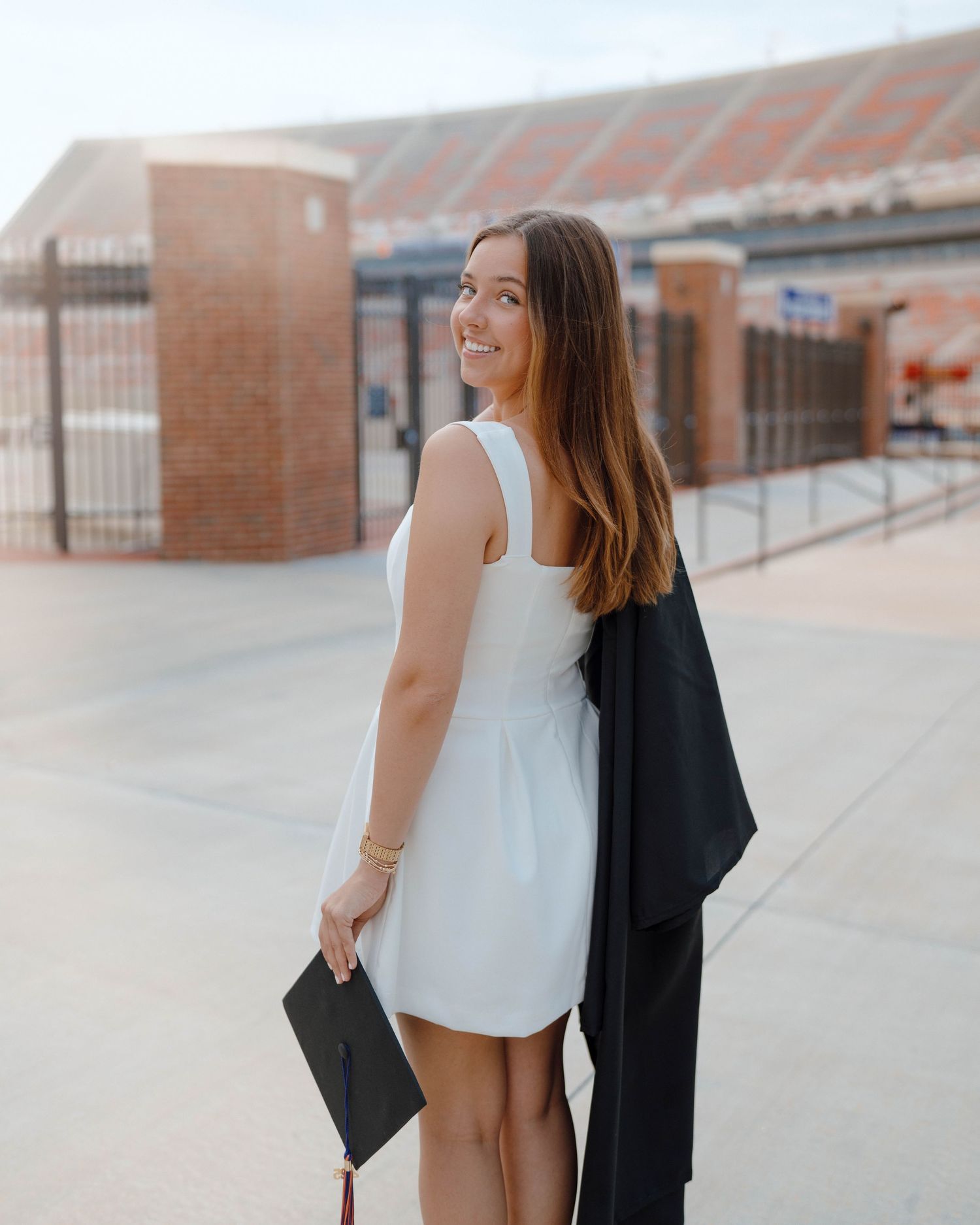A graduate in a white dress holds their graduation attire and diploma while smiling at a brick building stadium entrance.