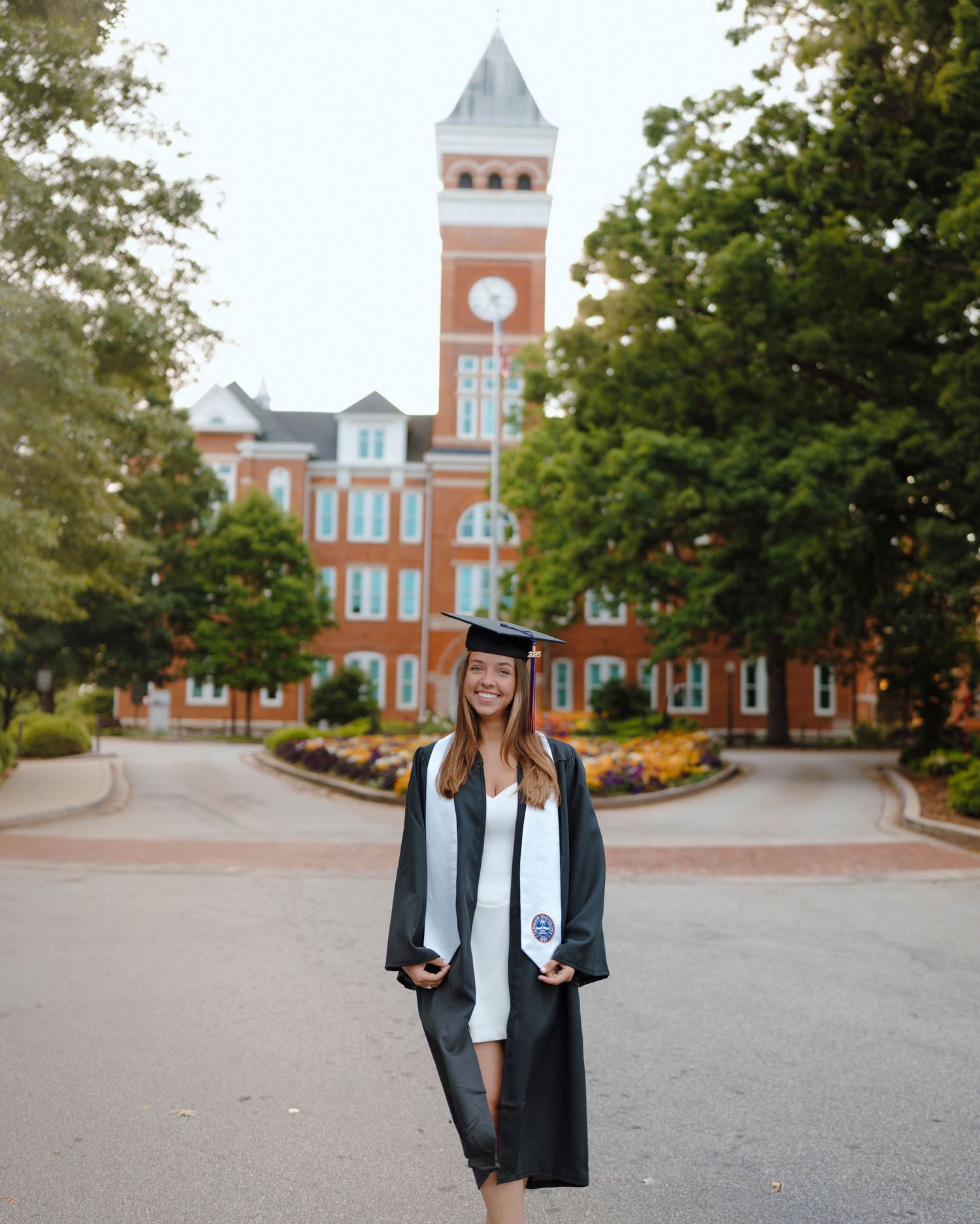 Graduate in cap and gown poses in front of iconic red brick university building with tall clock tower.