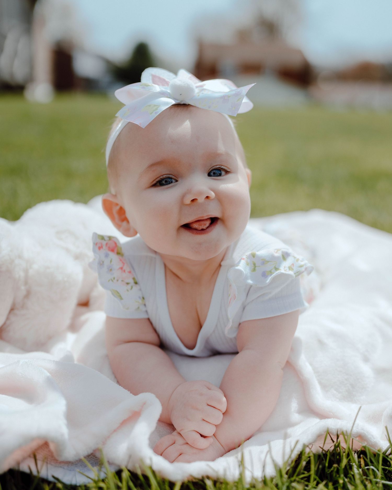 Smiling baby wearing white dress and bow lying on blanket outdoors in sunny garden setting.