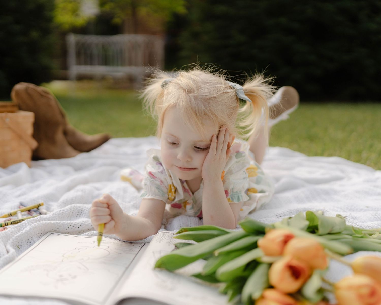 Child lying on blanket outdoors with orange tulips and book on sunny spring day.