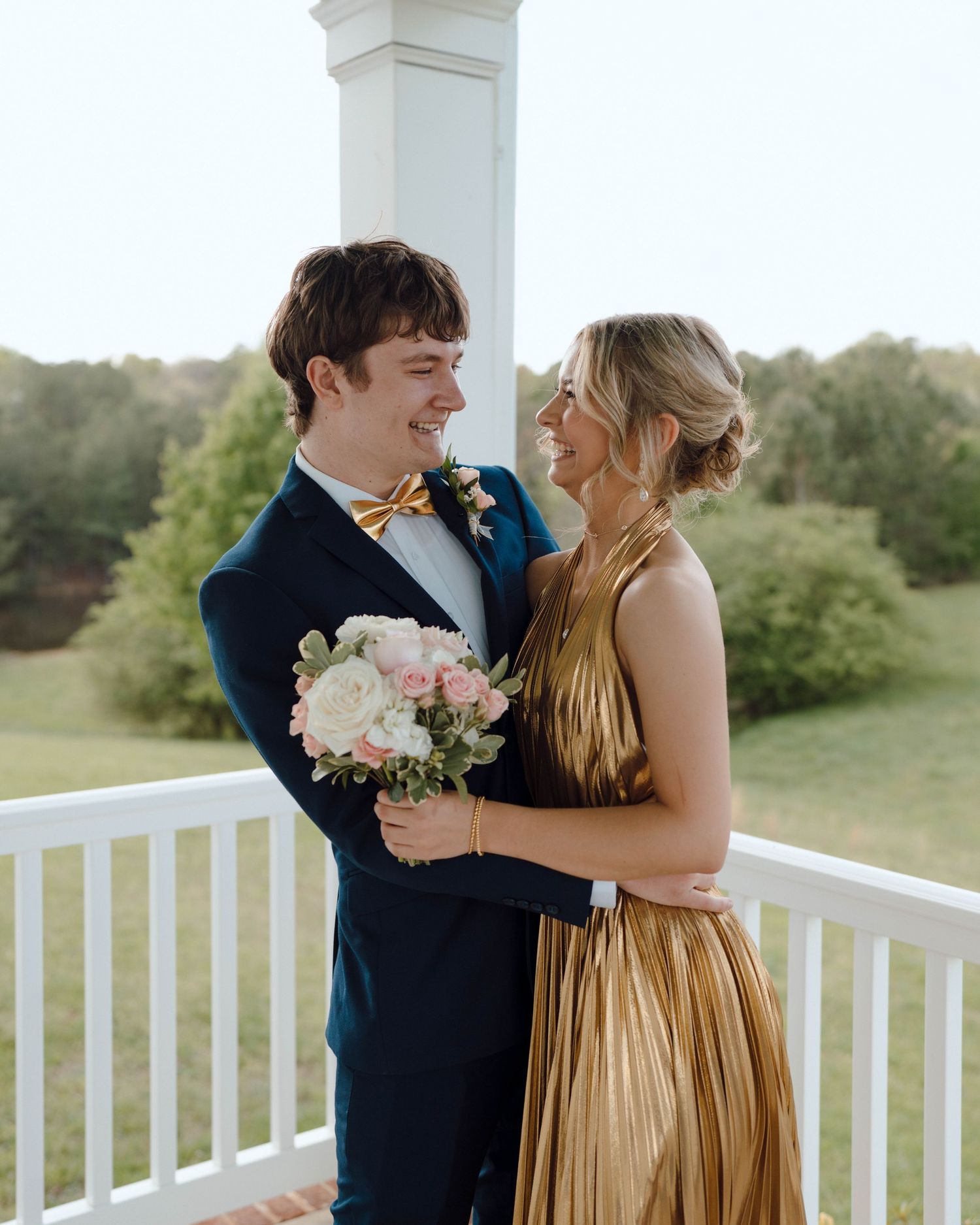A couple in formal attire share a romantic moment on a white porch, with the bride holding a pink and white bouquet.