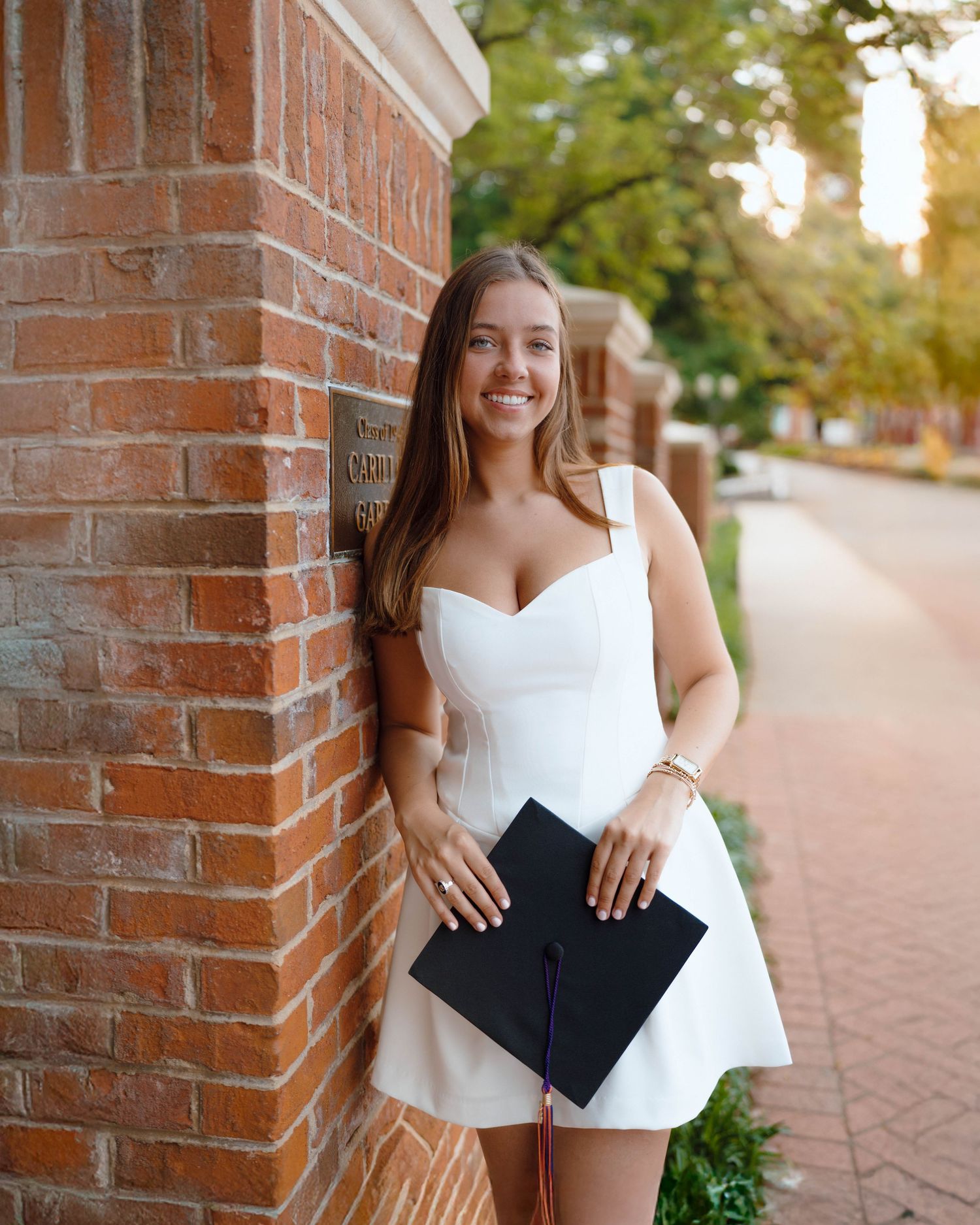 Graduate posing in white dress with diploma holder against brick column on tree-lined college campus at sunset.
