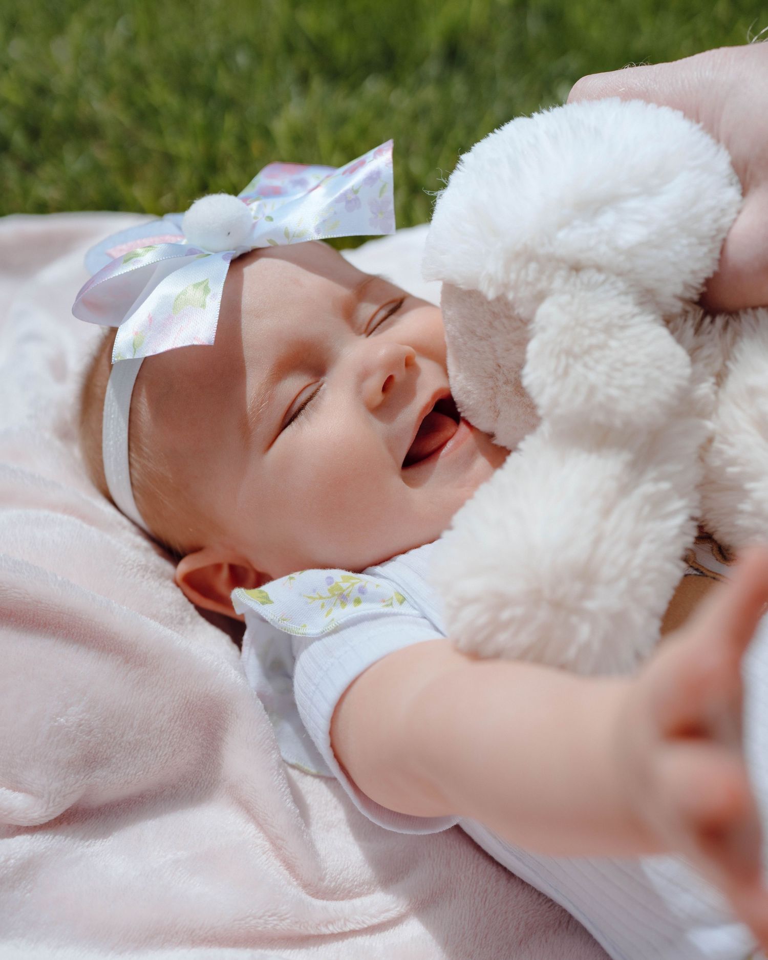 Laughing baby wearing white bow headband cuddles with plush toy on pink blanket.