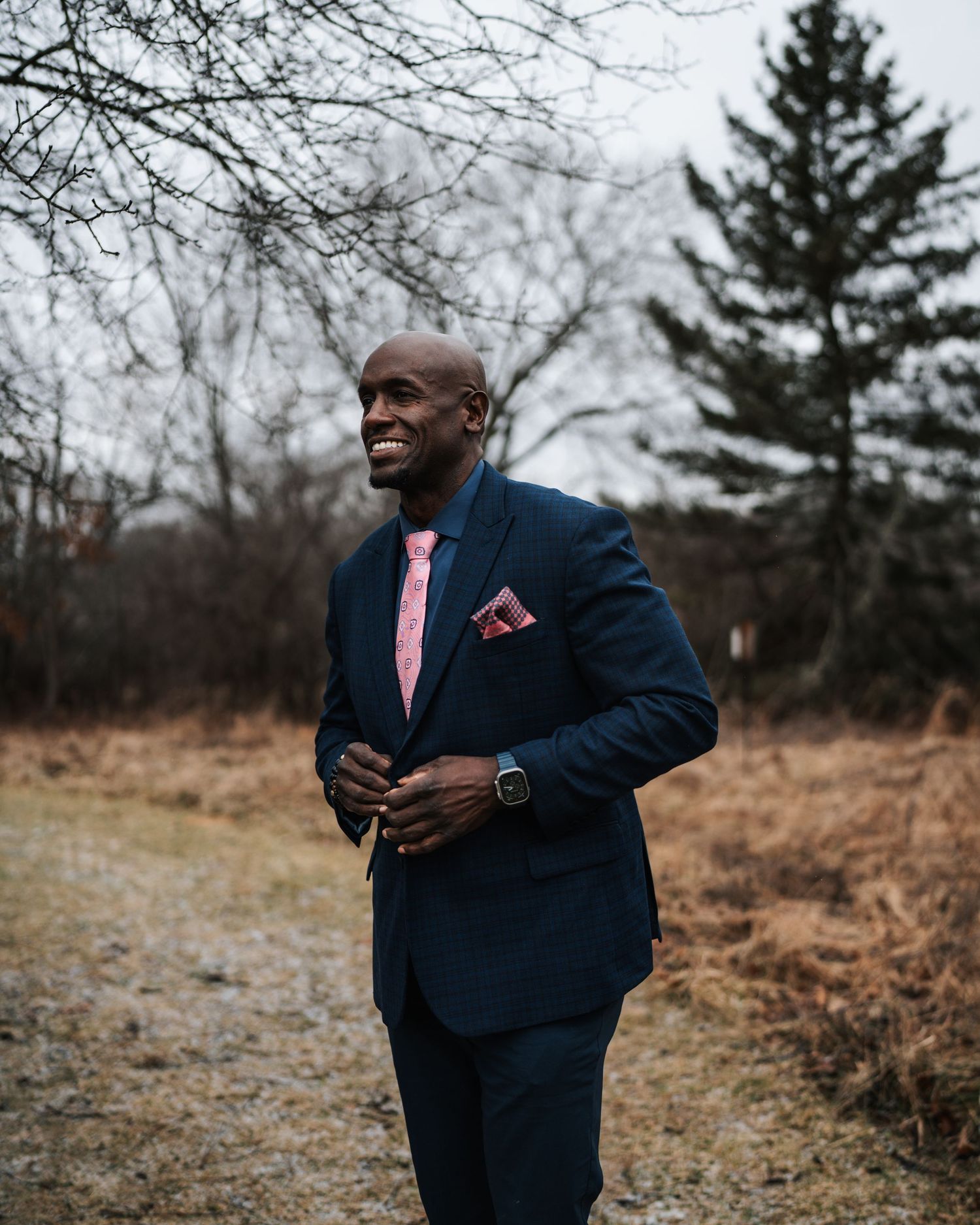 Professional in navy suit and pink tie poses outdoors in natural setting.