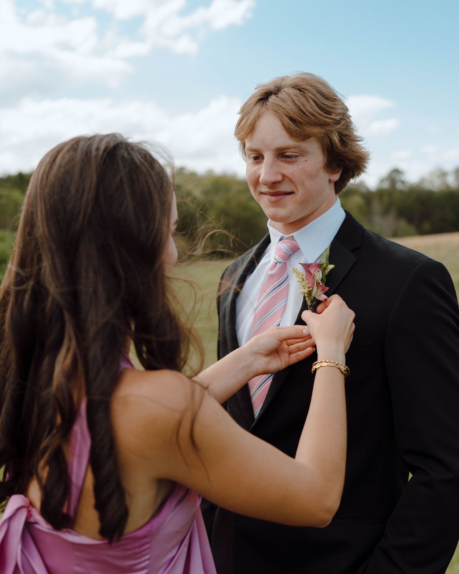 A couple share an intimate moment outdoors as someone adjusts a boutonniere on a black suit with pink tie.