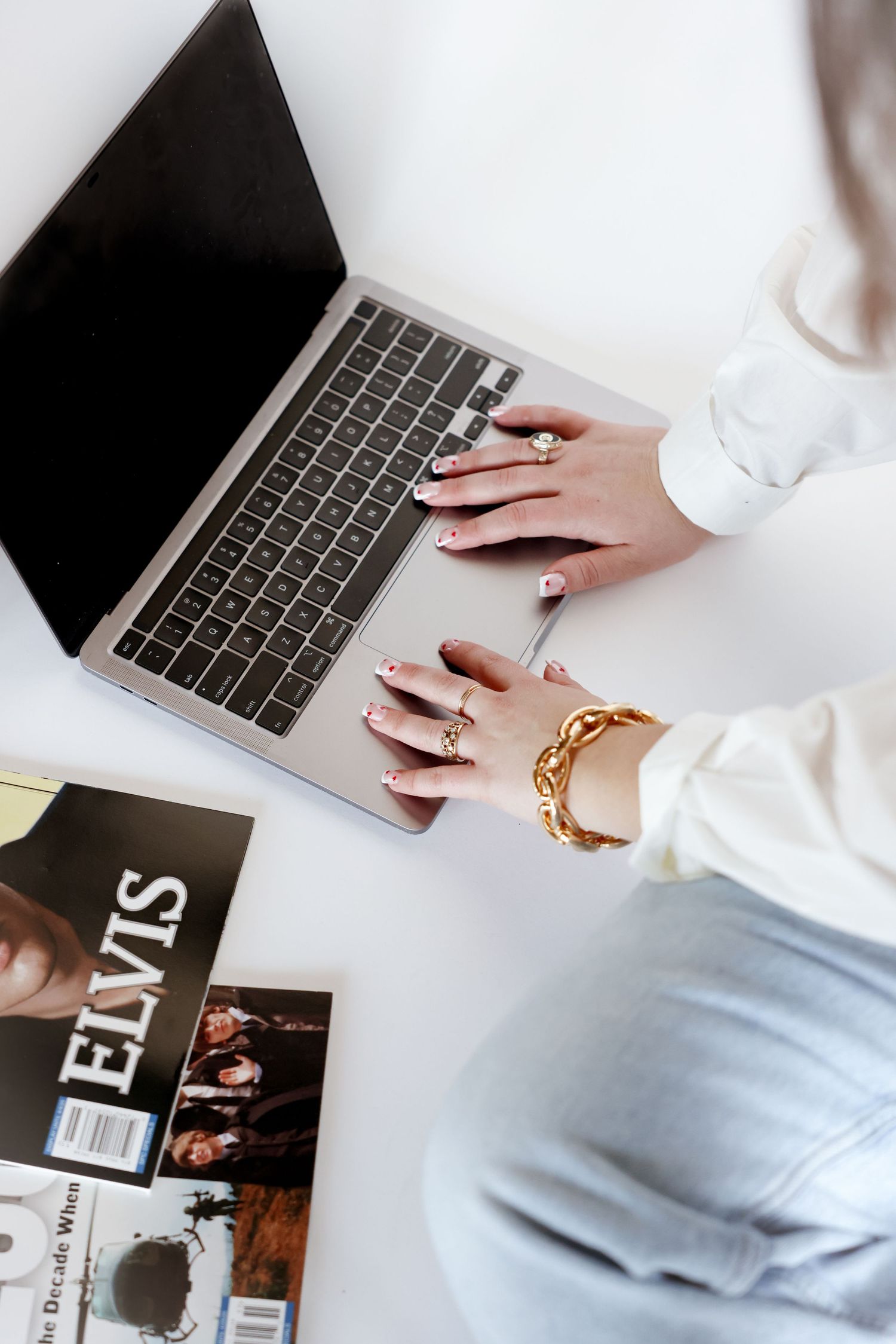 Hands with gold bracelet typing on laptop keyboard next to fashion magazine.