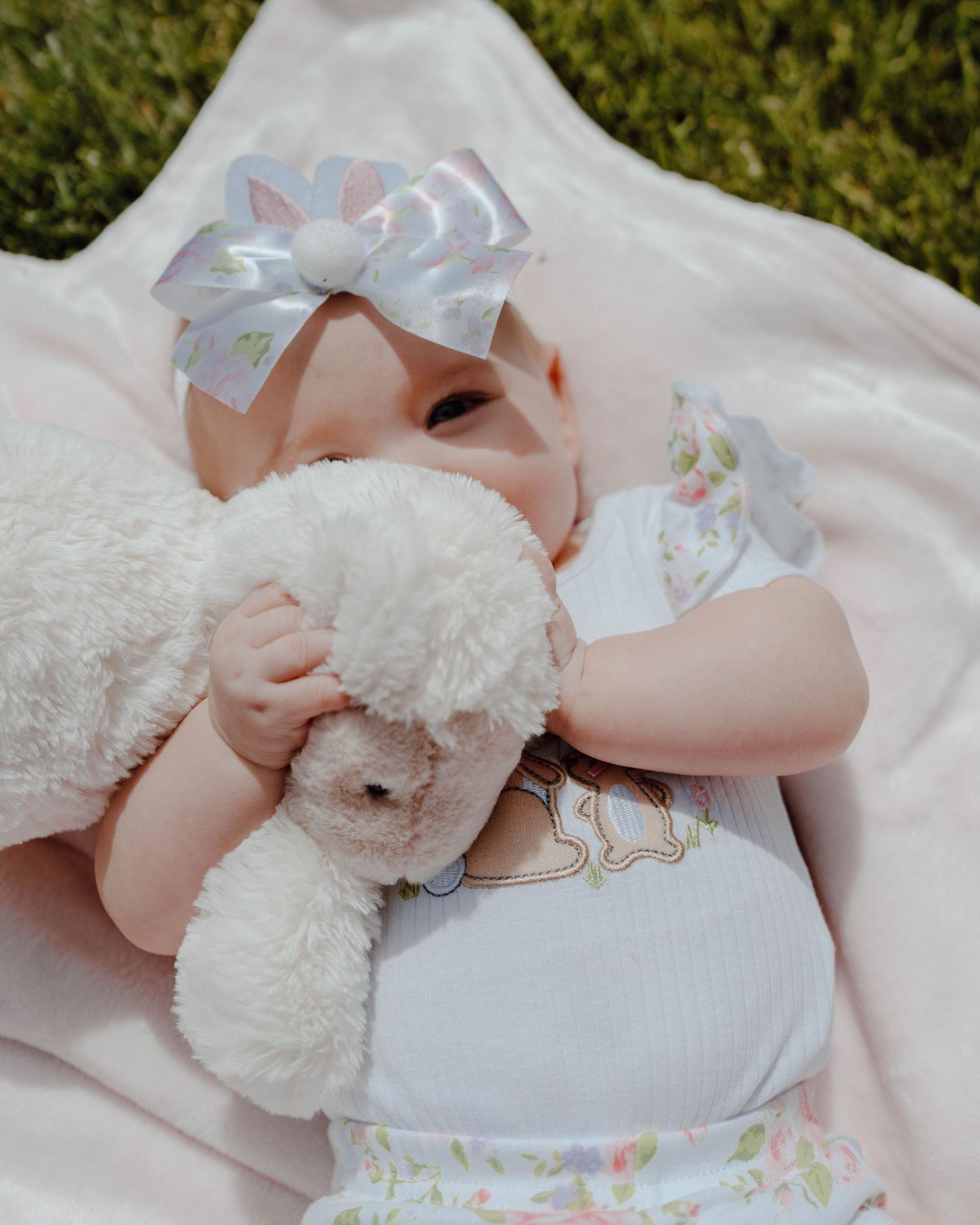 Baby in white dress with silver bow headband lying on pink blanket while holding plush white teddy bear.