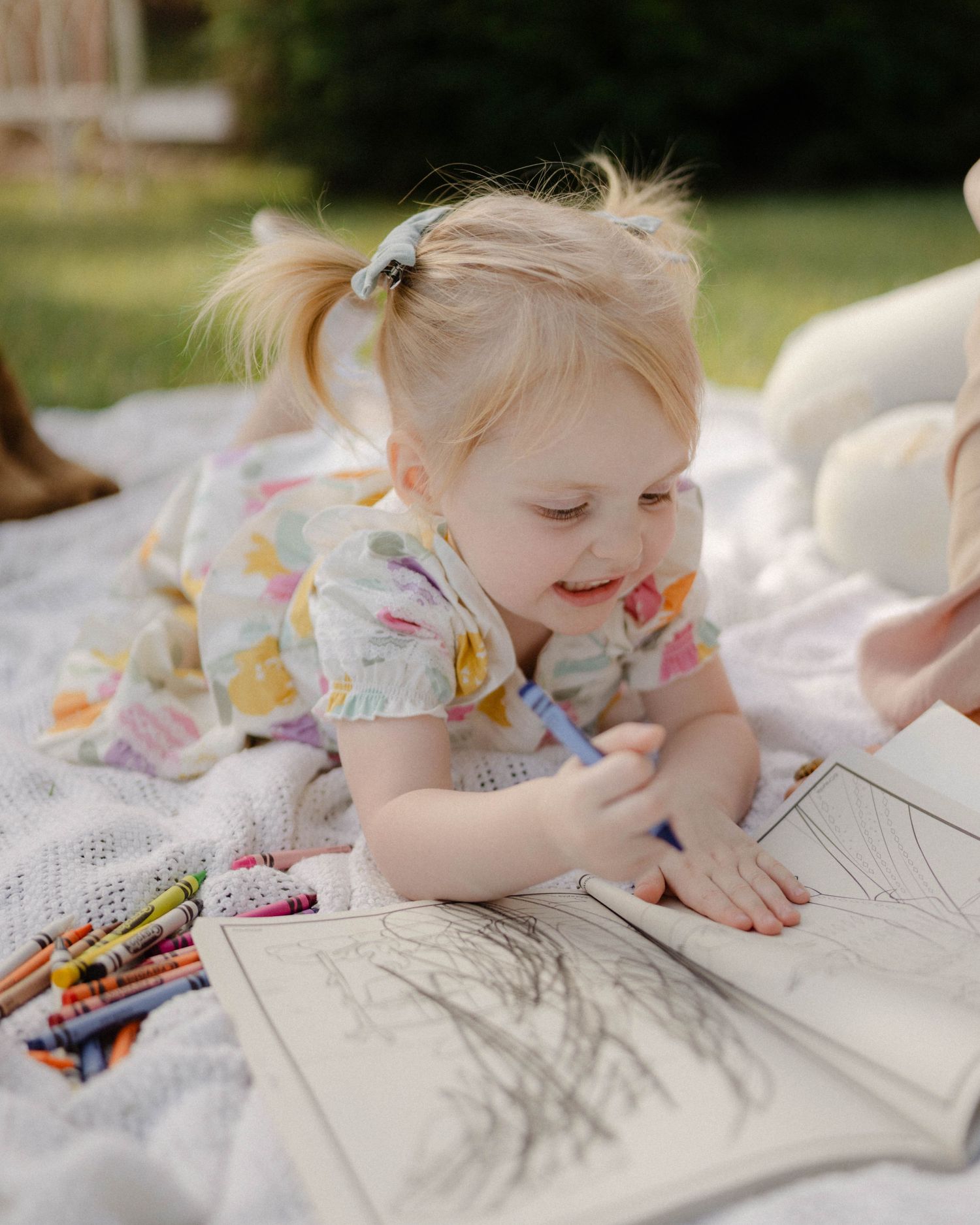 Child lies on a white blanket outdoors while coloring with crayons on paper during a sunny day.