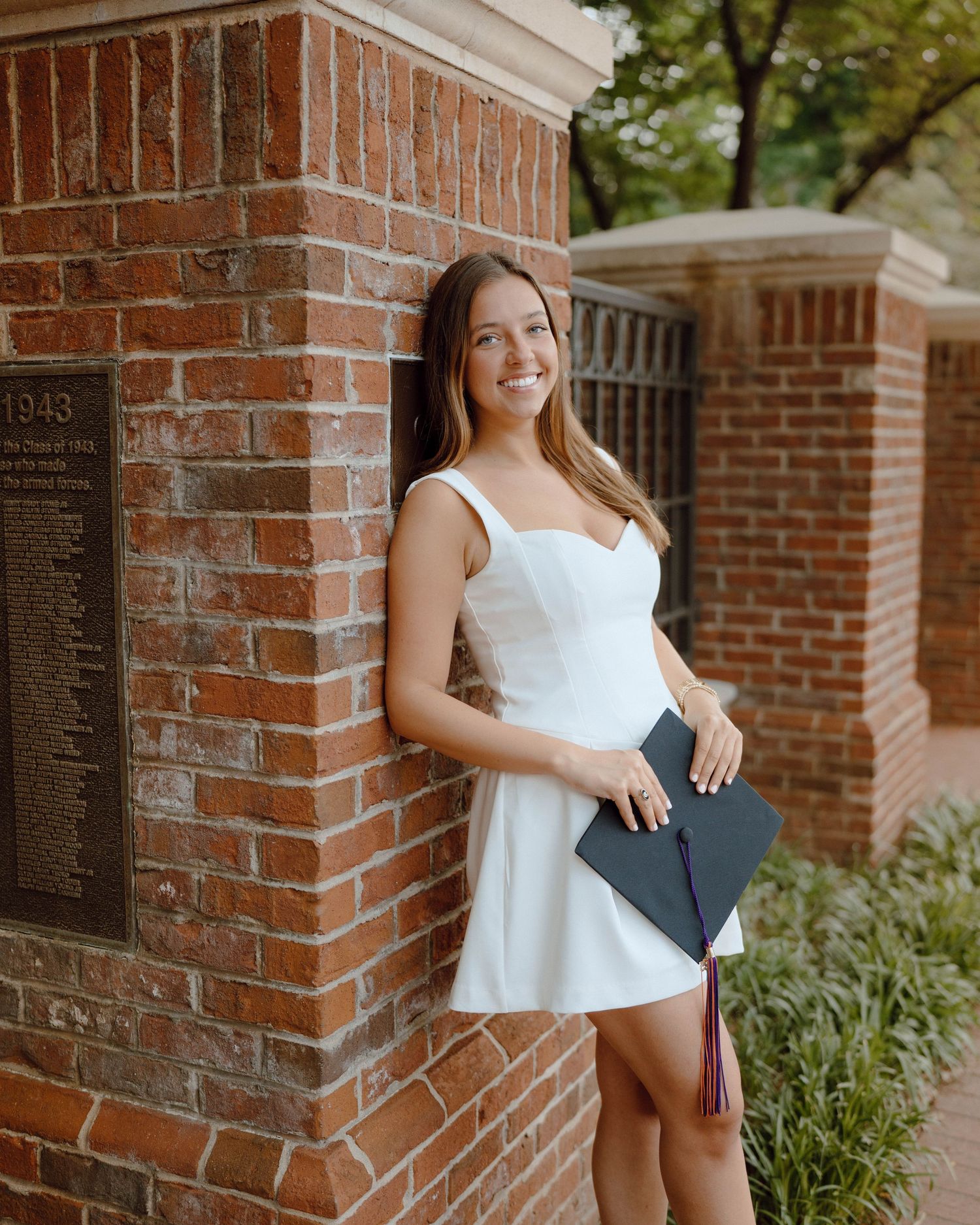 Graduate in a white dress holds a mortarboard while standing next to a brick column during a sunny day outside.