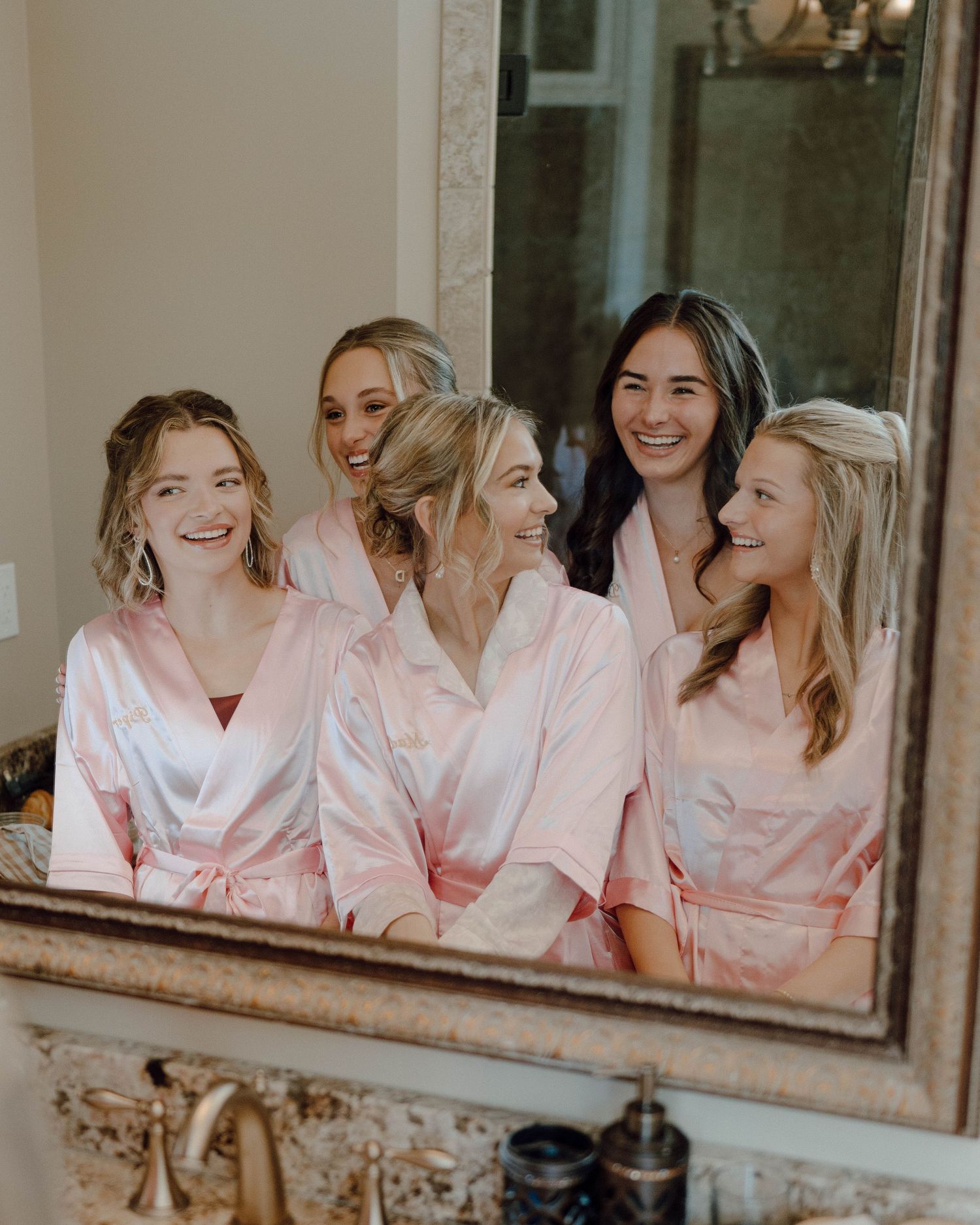 Group in matching pink satin robes smiling together in a bathroom mirror reflection on a special occasion.