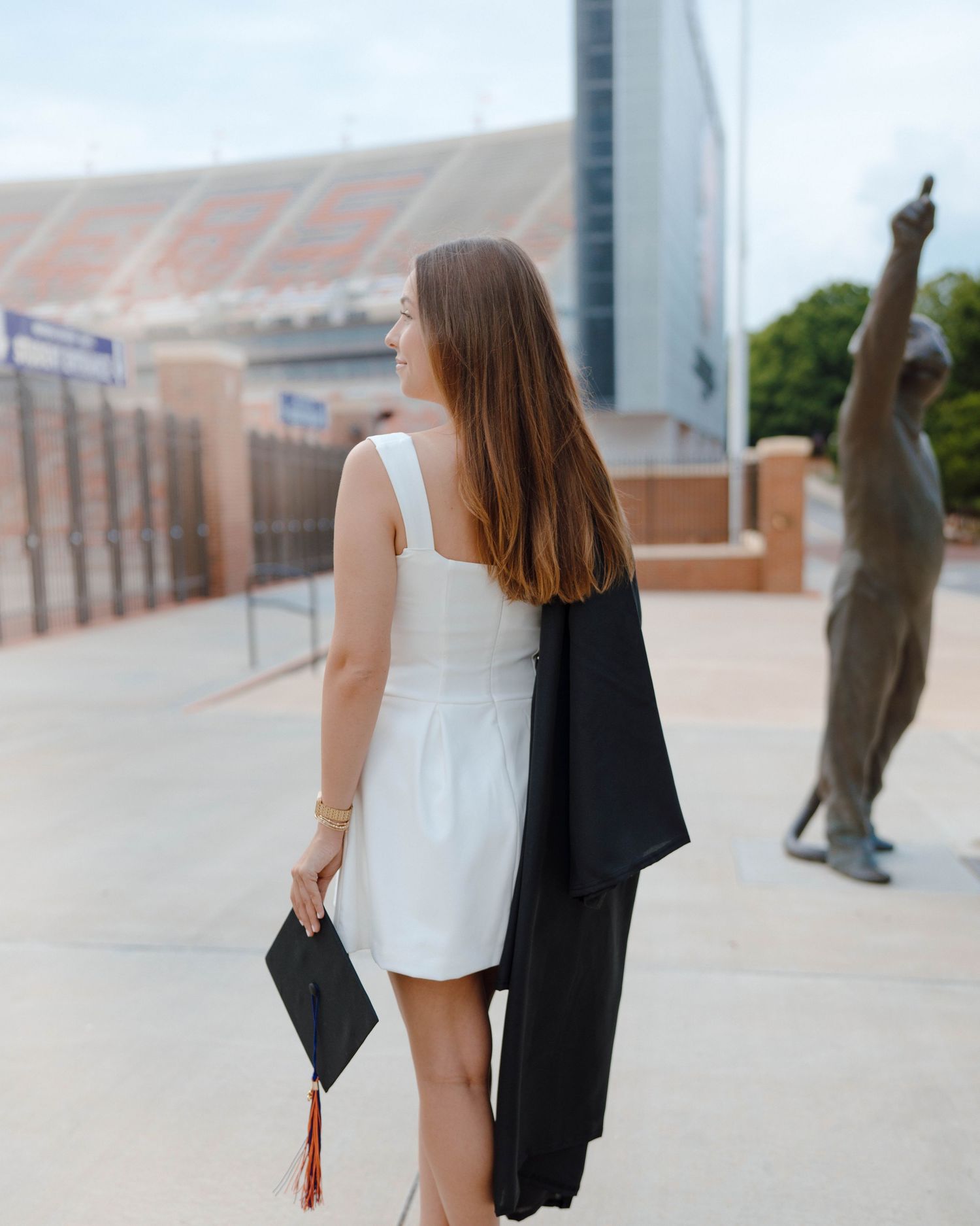 Graduate in white dress holding diploma case and gown stands near bronze statue outside college stadium.
