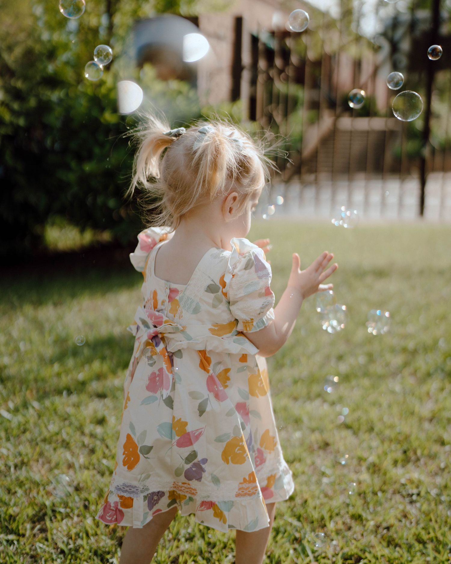 A young child in a floral dress plays with bubbles in a sunny backyard garden.