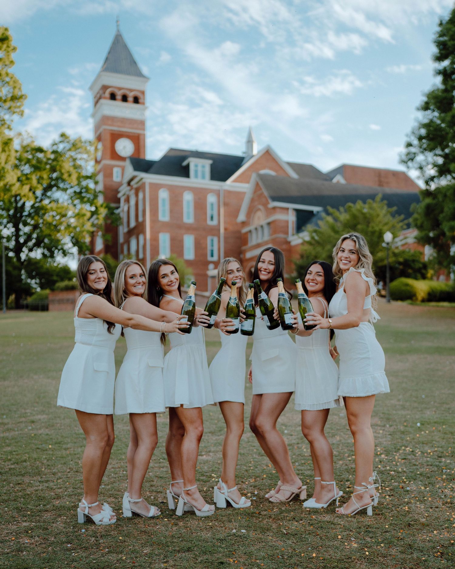 Friends in coordinated white dresses pose with drinks on a lawn in front of a brick university building with tower.