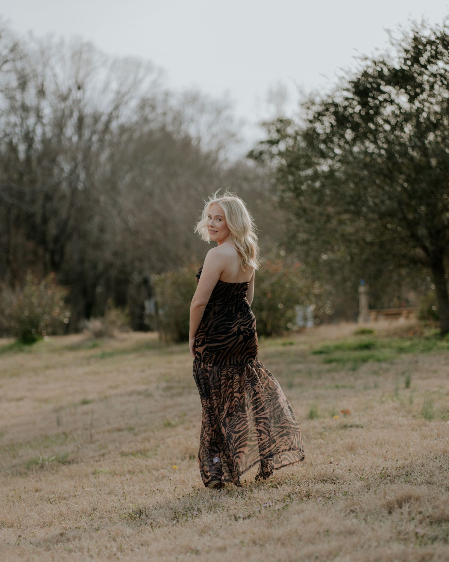 A person in a flowing dress stands in a rustic outdoor setting during golden hour with trees in the background.
