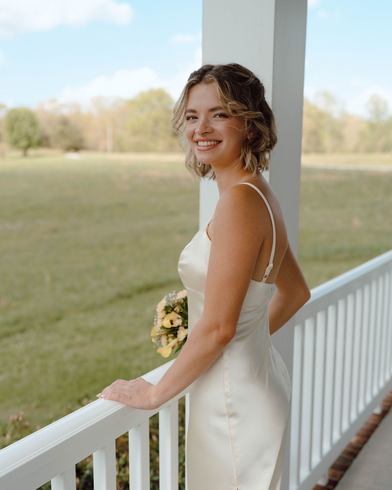 A person in a white formal dress stands smiling on a white porch railing overlooking a grassy field.