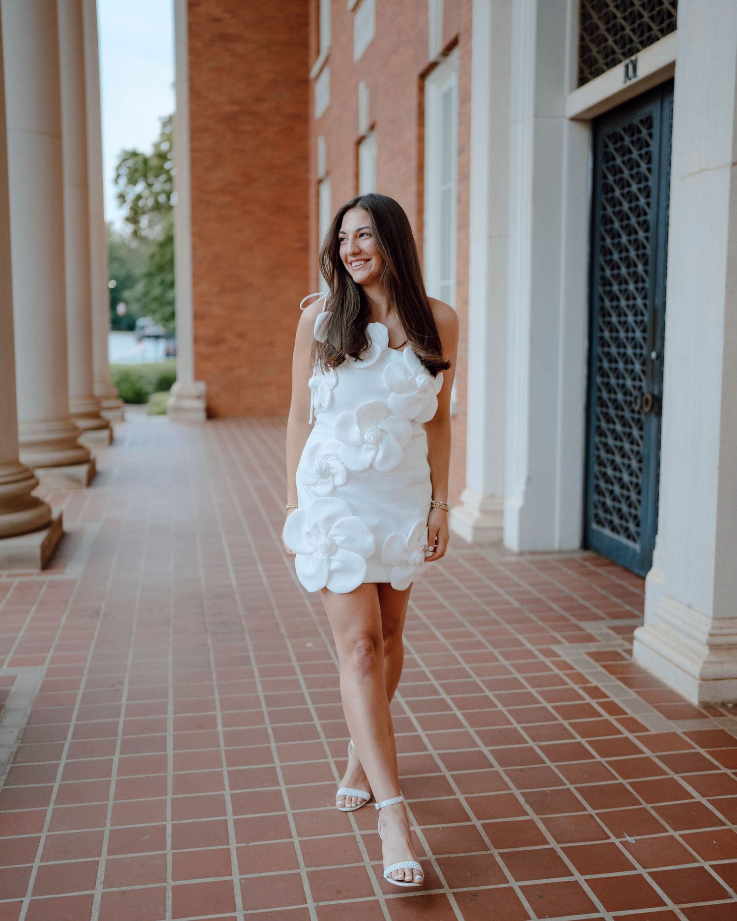 Person in white ruffled mini dress and sandals poses elegantly on a brick walkway near classic columns.