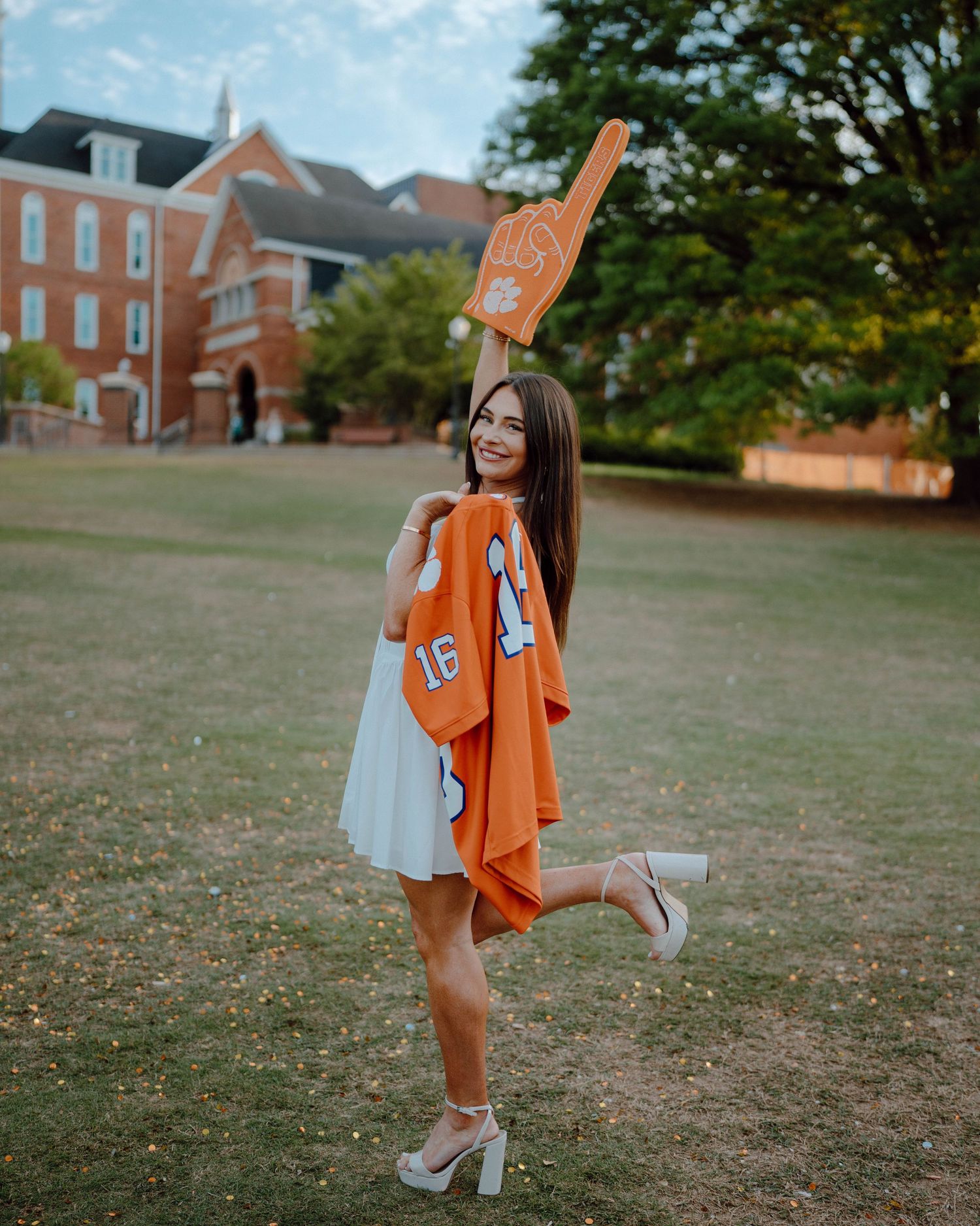 A person in a white dress poses on a college campus lawn while holding an orange jersey and number foam finger.