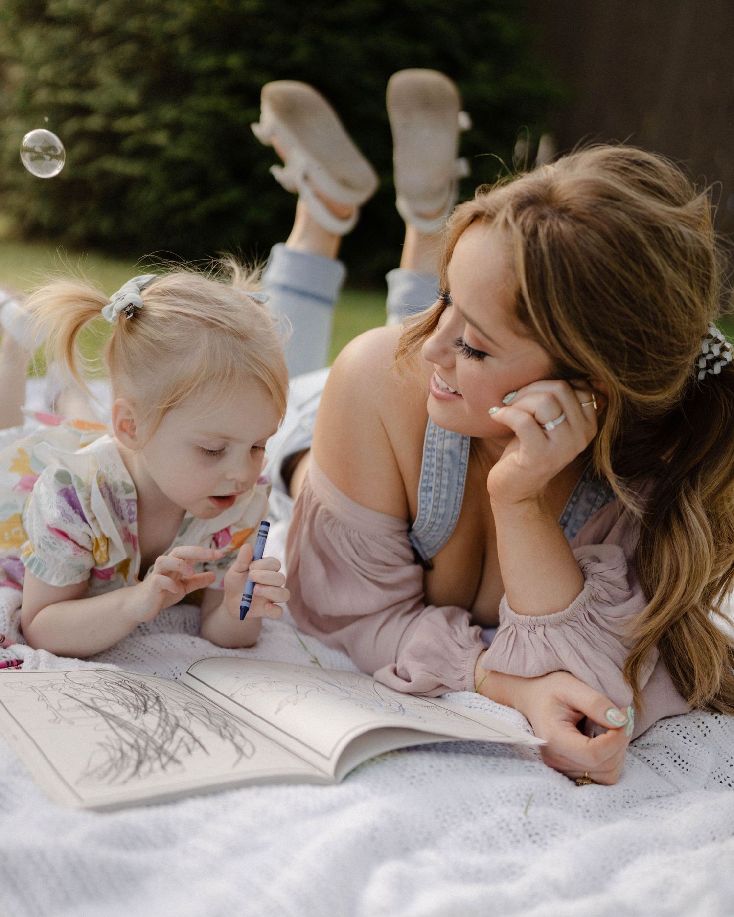 Mother and daughter lie on a blanket outdoors reading and coloring together on a relaxing afternoon.