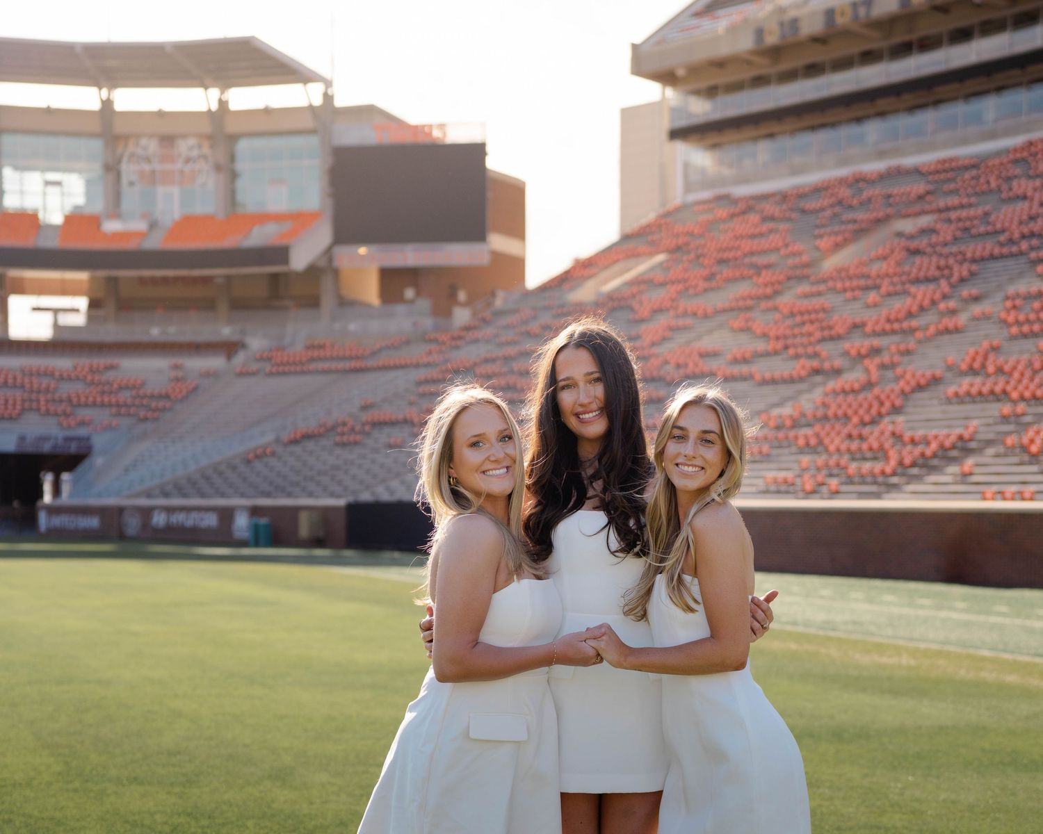 Three people in white dresses pose together on a football field in front of stadium seating at sunset.