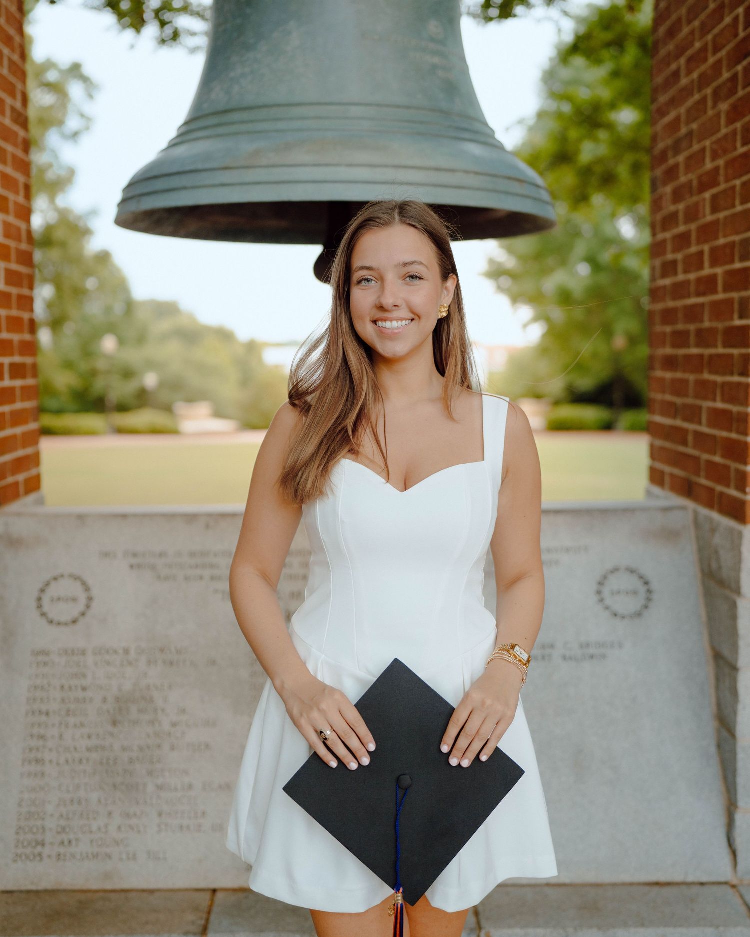 Graduate in white dress holding diploma while standing under large bell at college memorial wall.