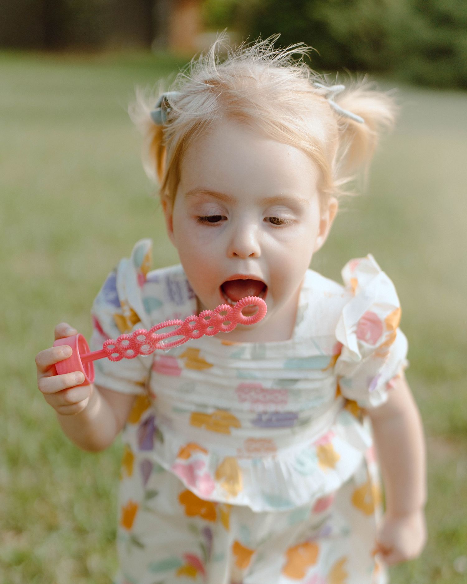A small child in a floral dress blows a pink plastic bubble wand while playing outdoors on a sunny day.