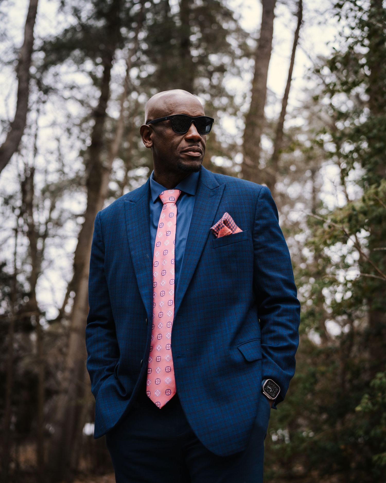 Stylishly dressed person in a navy blazer and coral tie poses confidently against a wooded backdrop.