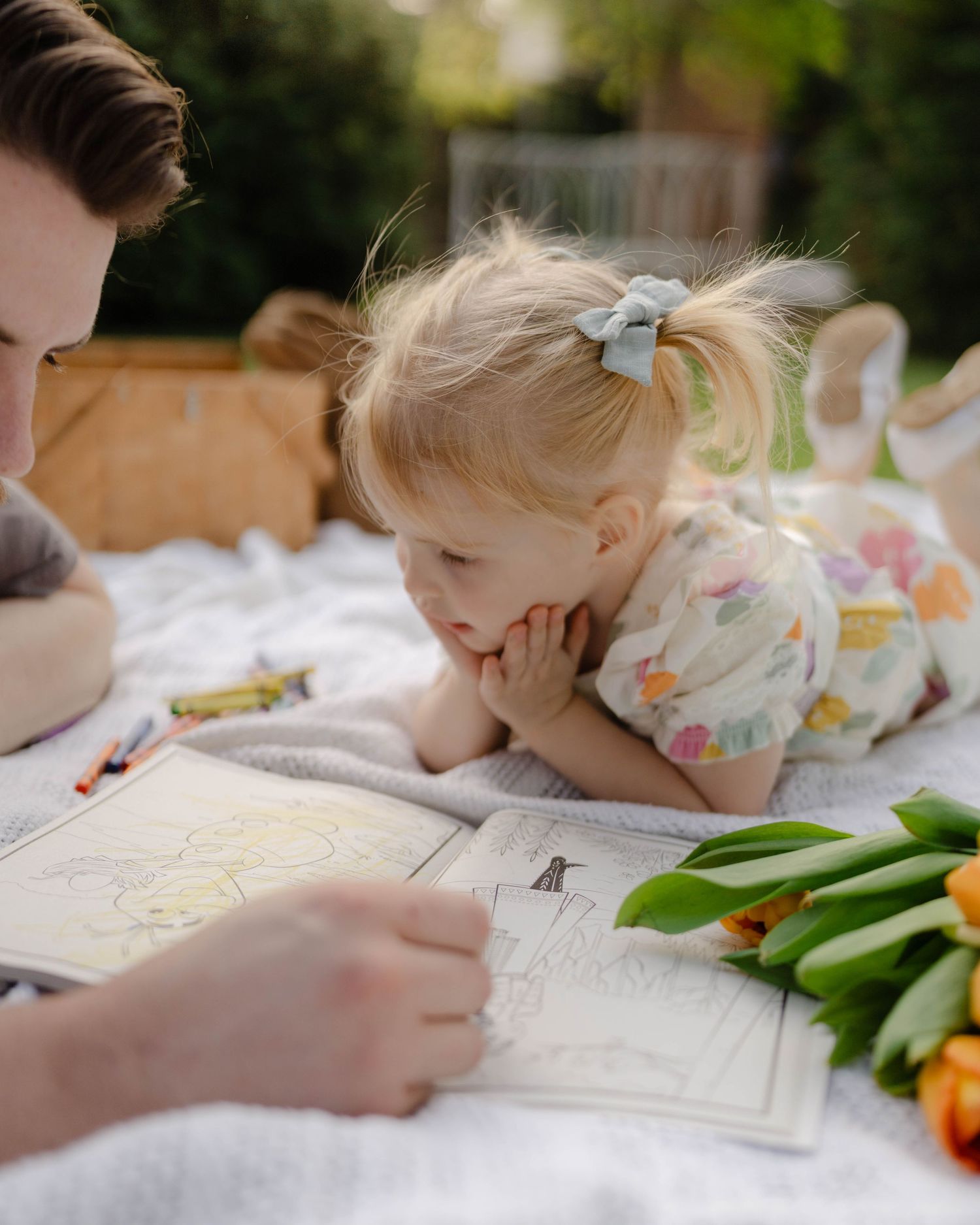 Small child lays on picnic blanket looking at book with flowers and writing materials beside them.
