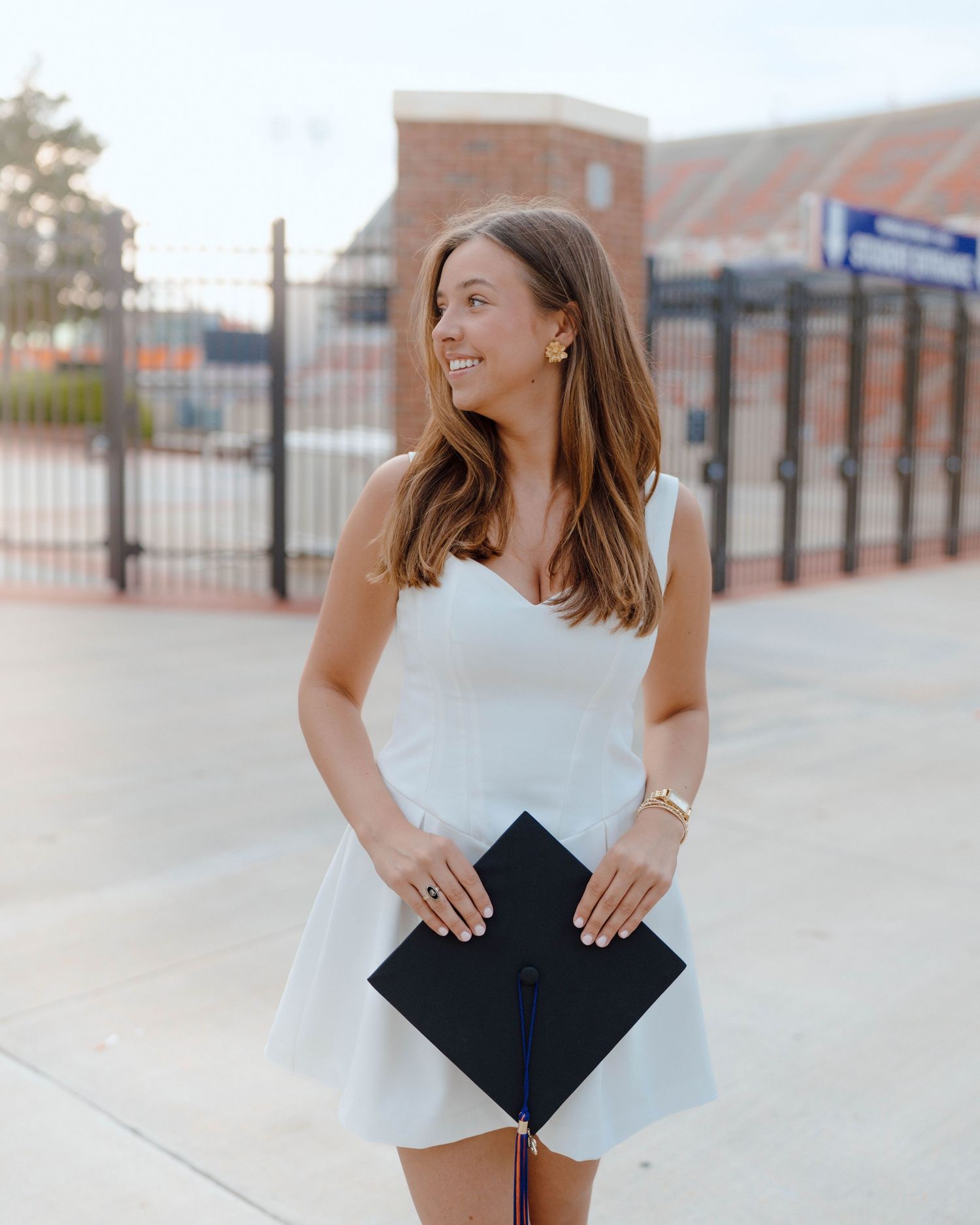 A woman in a white dress holds a graduation diploma outdoors on a campus plaza smiling to the side.