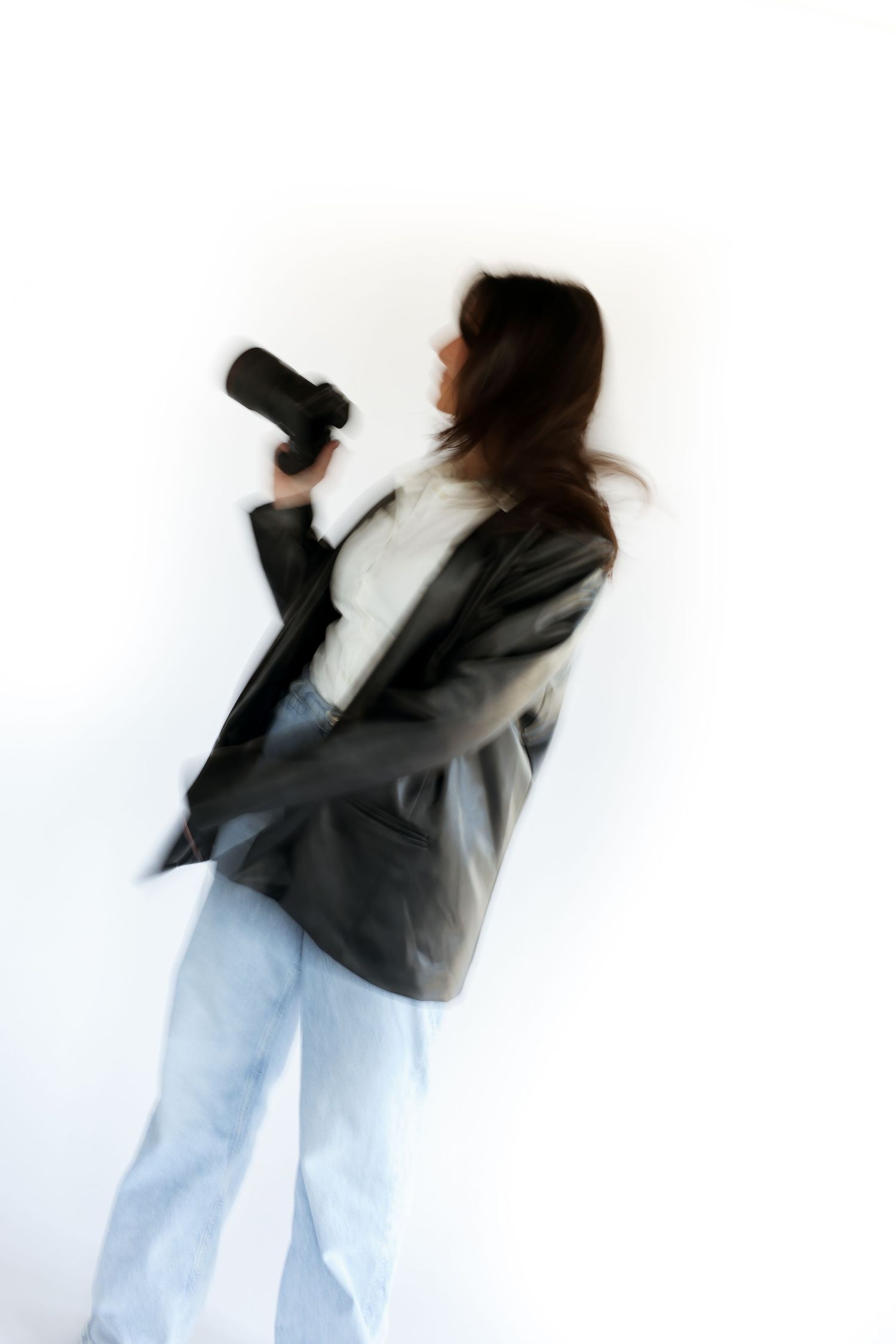 A person wearing a blazer and jeans stands sideways while holding a professional camera against a white background.