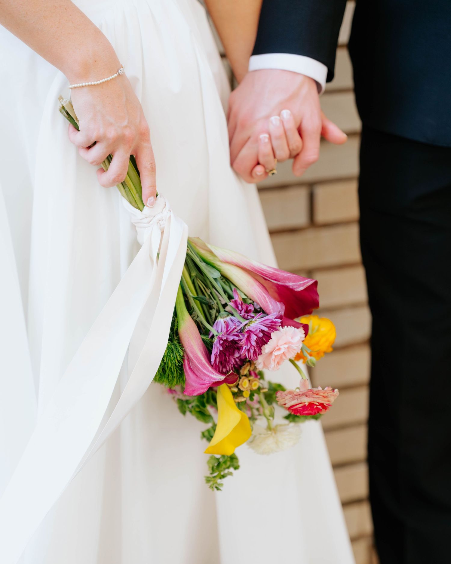 Bride holding colorful wedding bouquet with pink, yellow, and purple flowers while holding groom's hand in navy suit.
