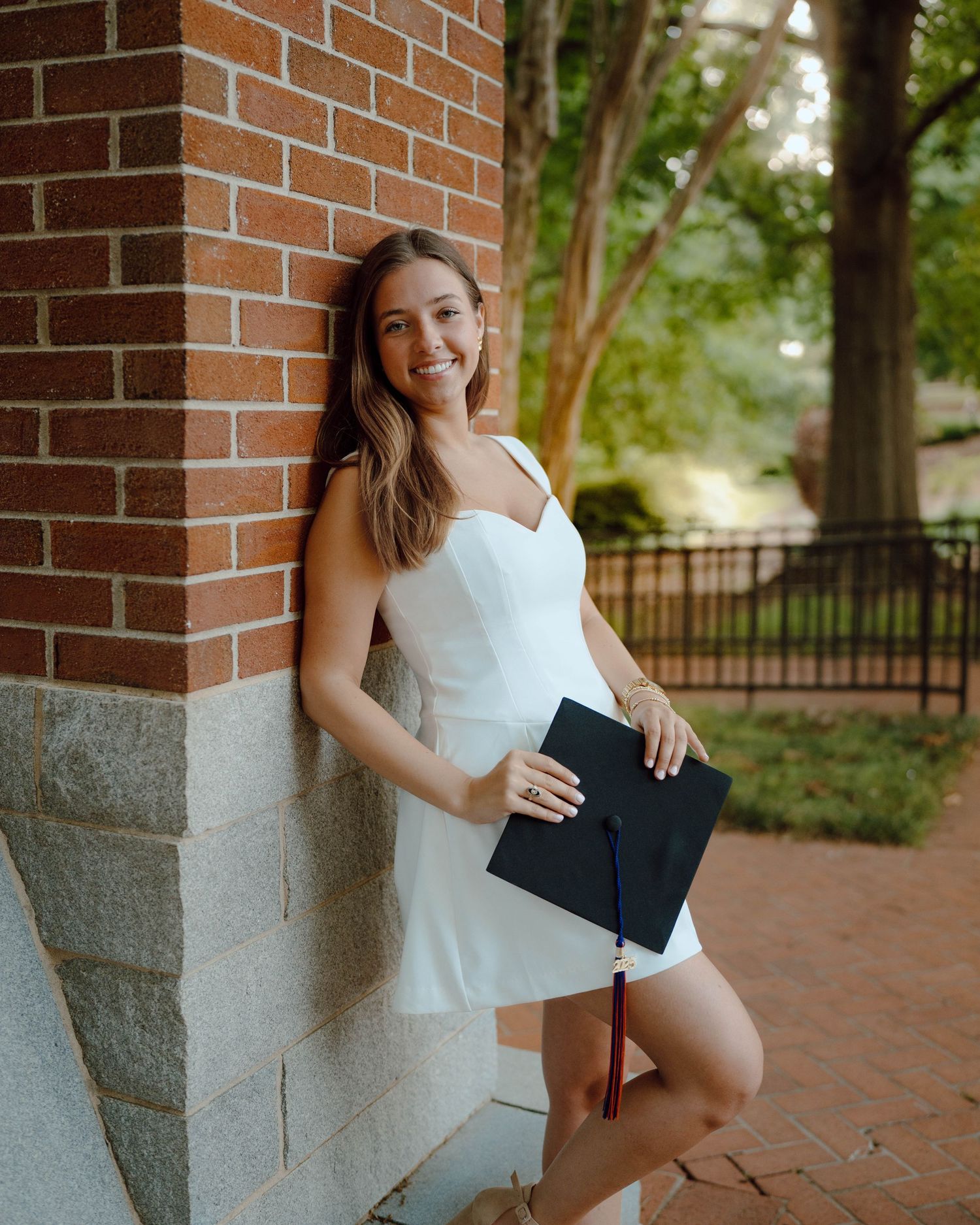 A graduate in a white dress poses with diploma against a brick column on a brick pathway surrounded by trees.