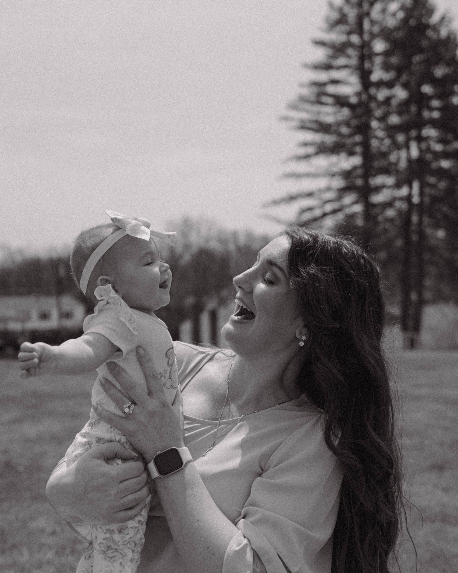 A parent gently lifts a smiling baby outdoors against a backdrop of evergreen trees in black and white photography.