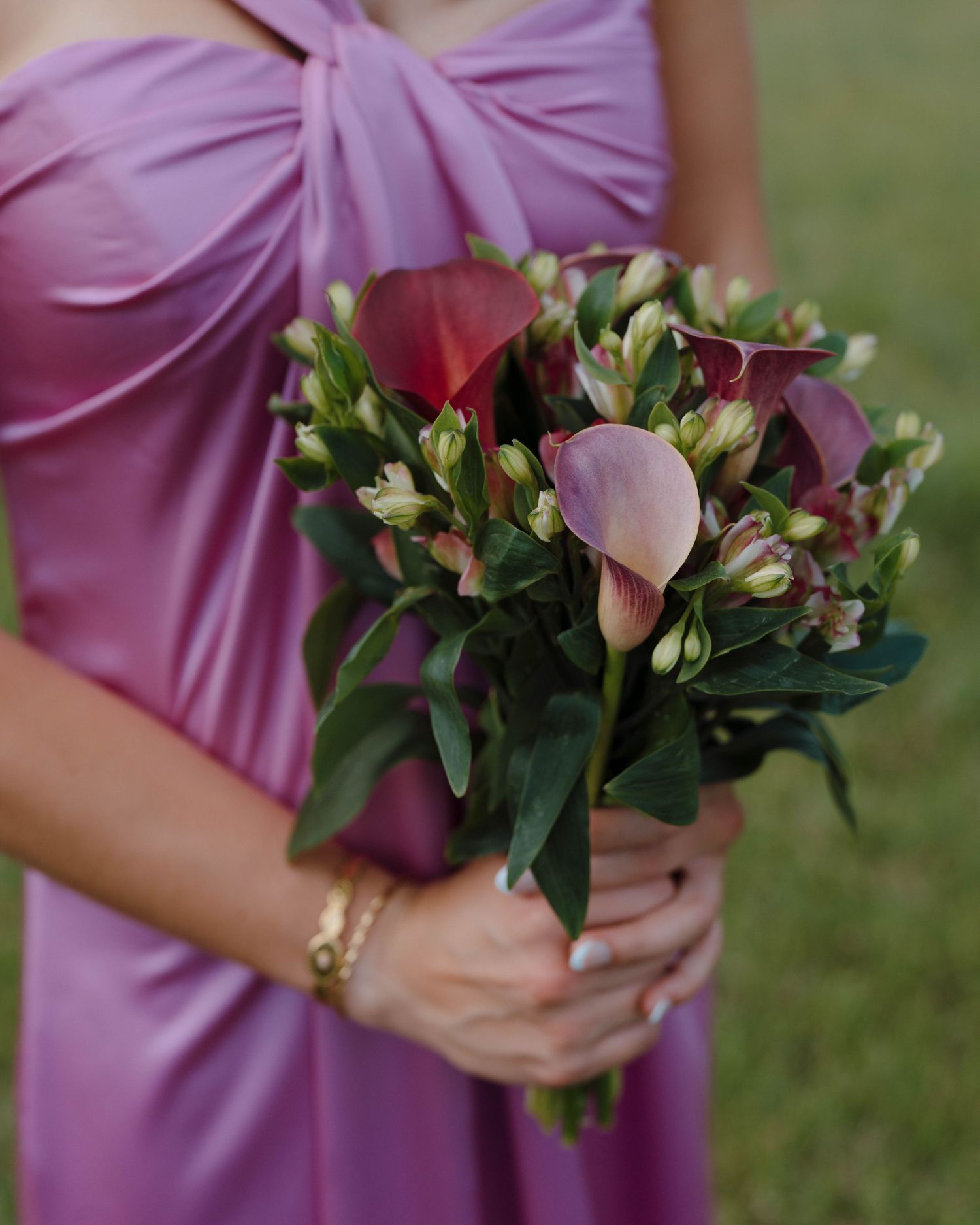 Close up of hands holding a wedding bouquet with burgundy calla lilies and pink flowers against a pink fabric dress.