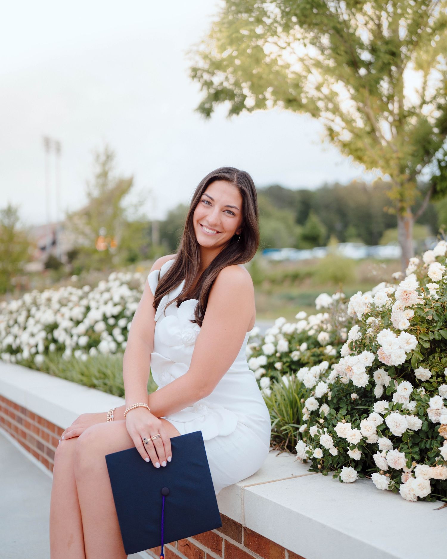 Someone poses on a brick ledge in a white dress with a navy blue clutch purse surrounded by blooming white flowers.
