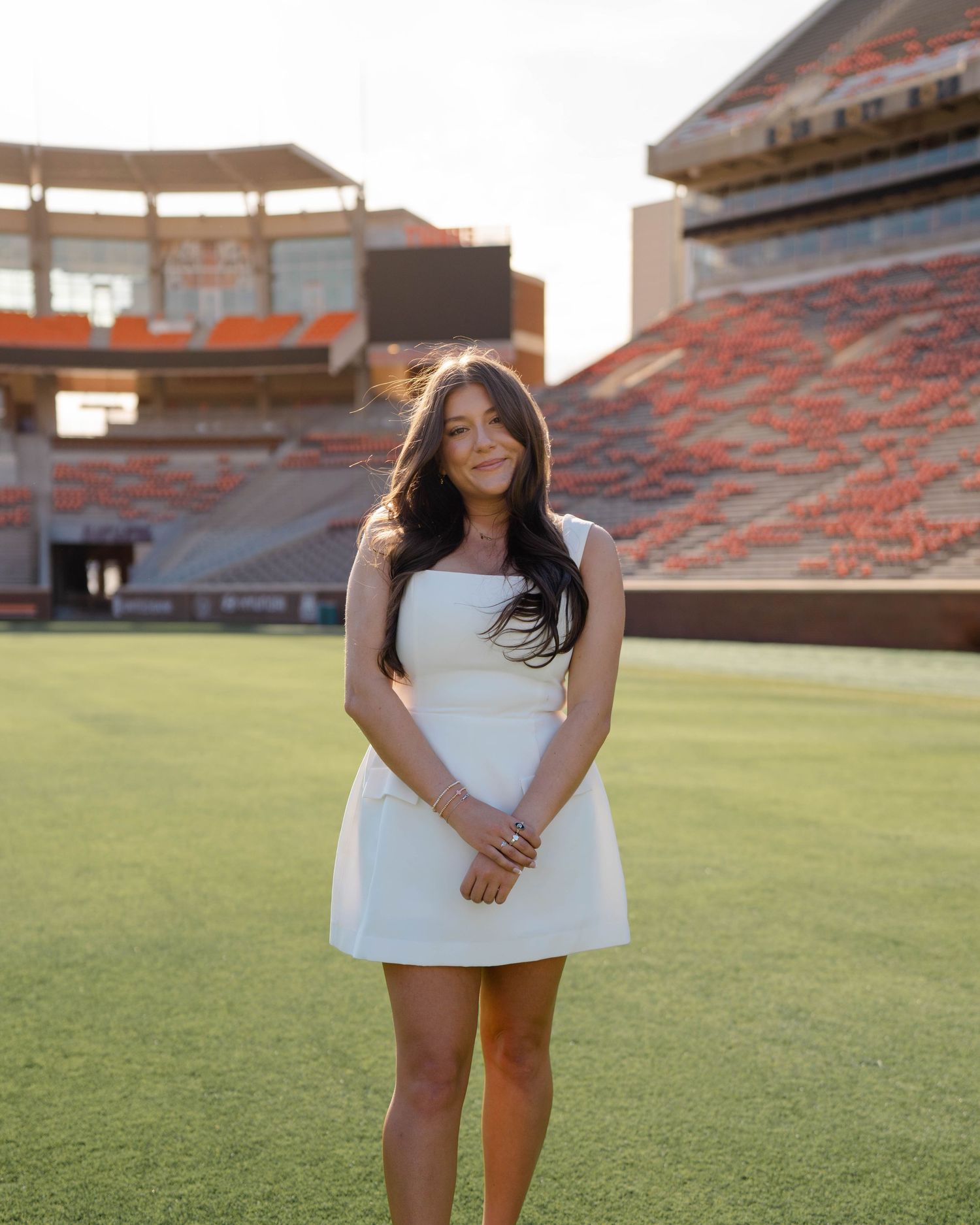 A person in a white dress stands on the green field of a college football stadium during golden hour.