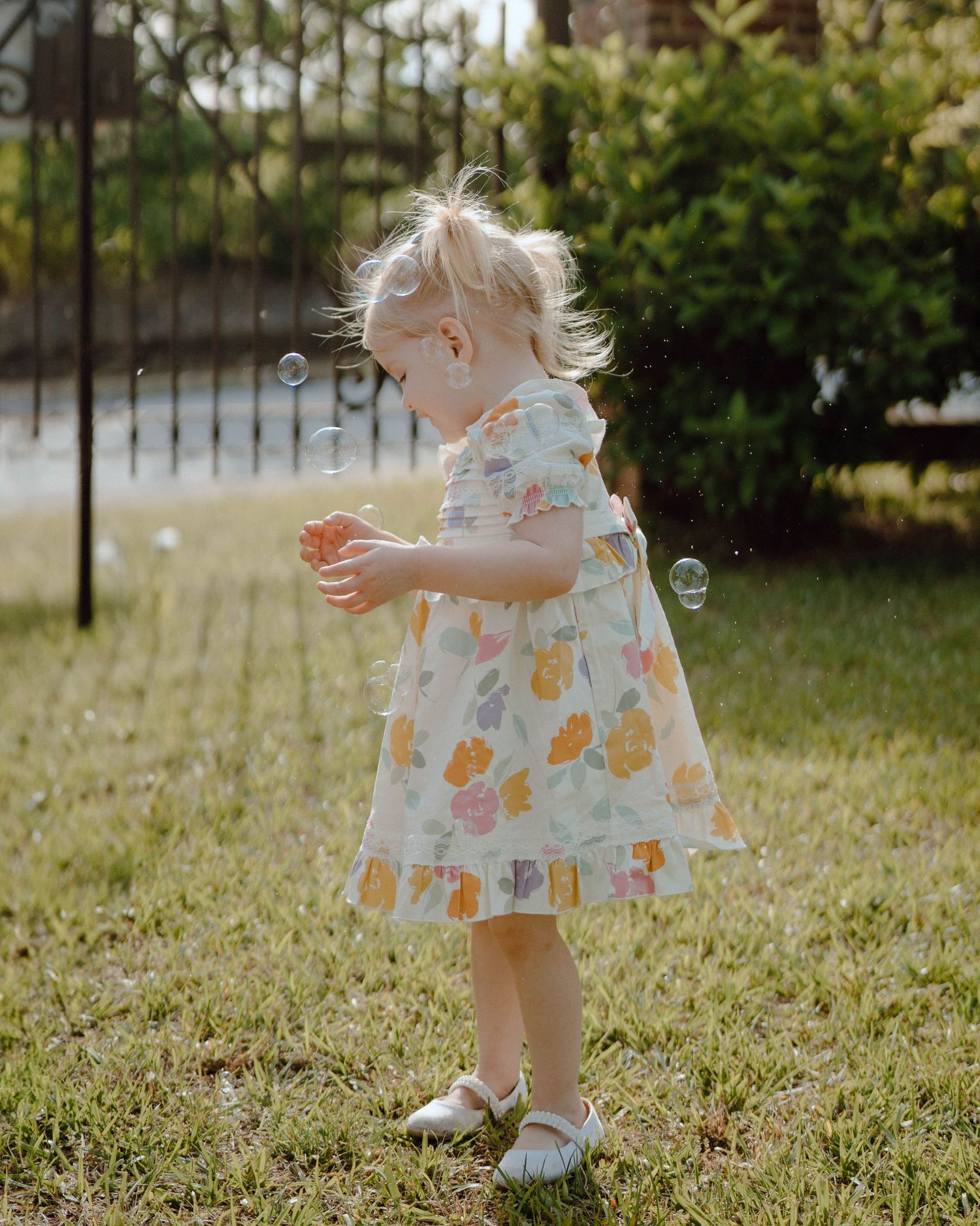 A toddler in a floral dress plays with bubbles in a sunlit garden setting.