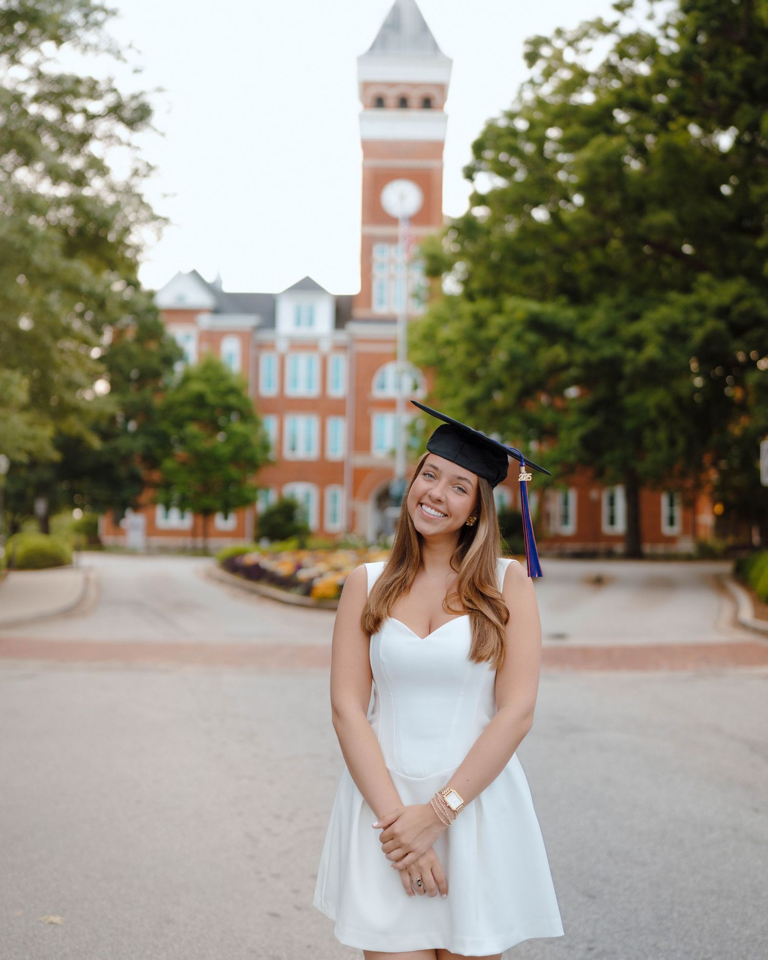 Graduate in white dress poses in front of red brick university building with prominent clock tower.