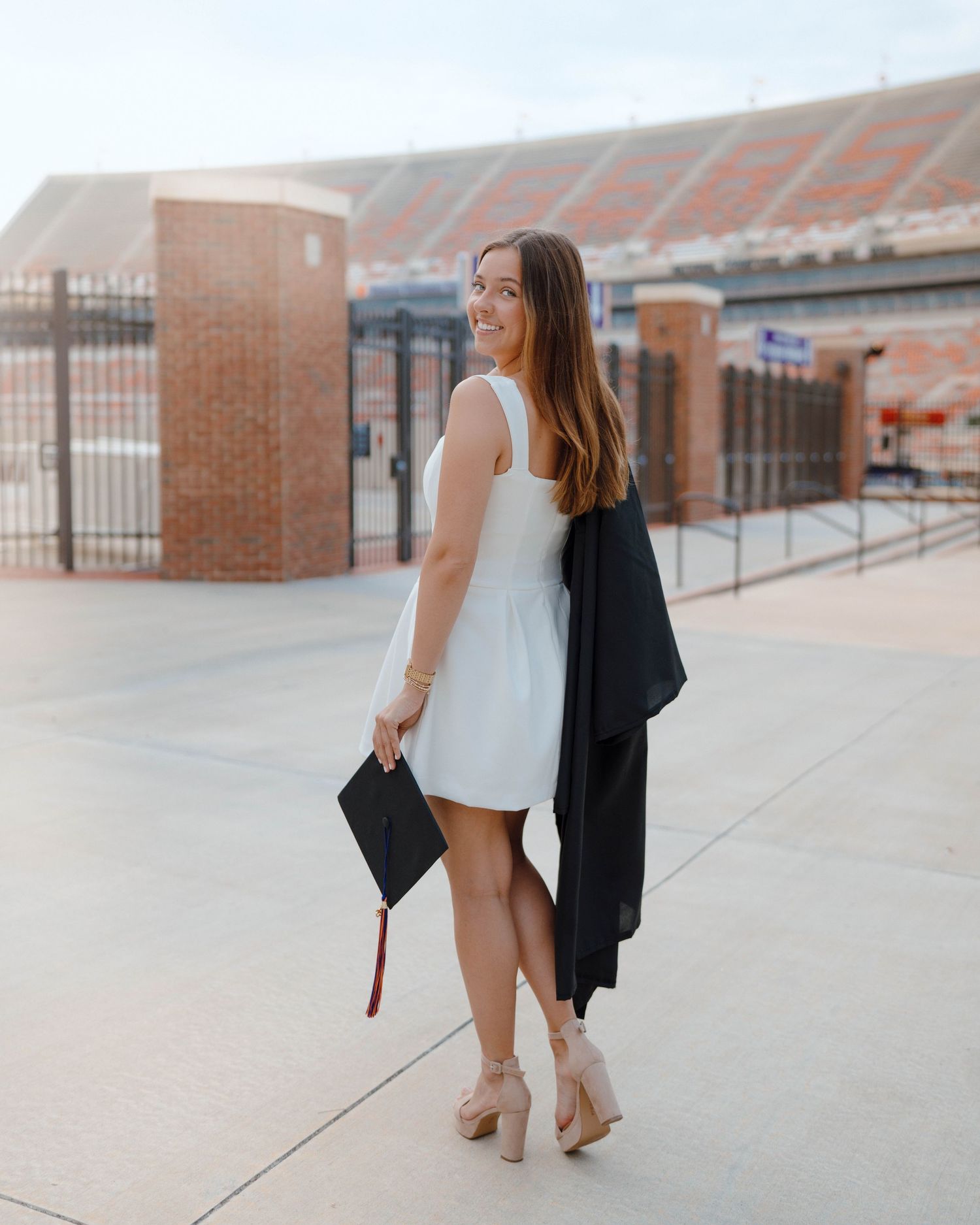 Graduate in a white dress and high heels stands with cap and gown draped over arm outside stadium on graduation day.