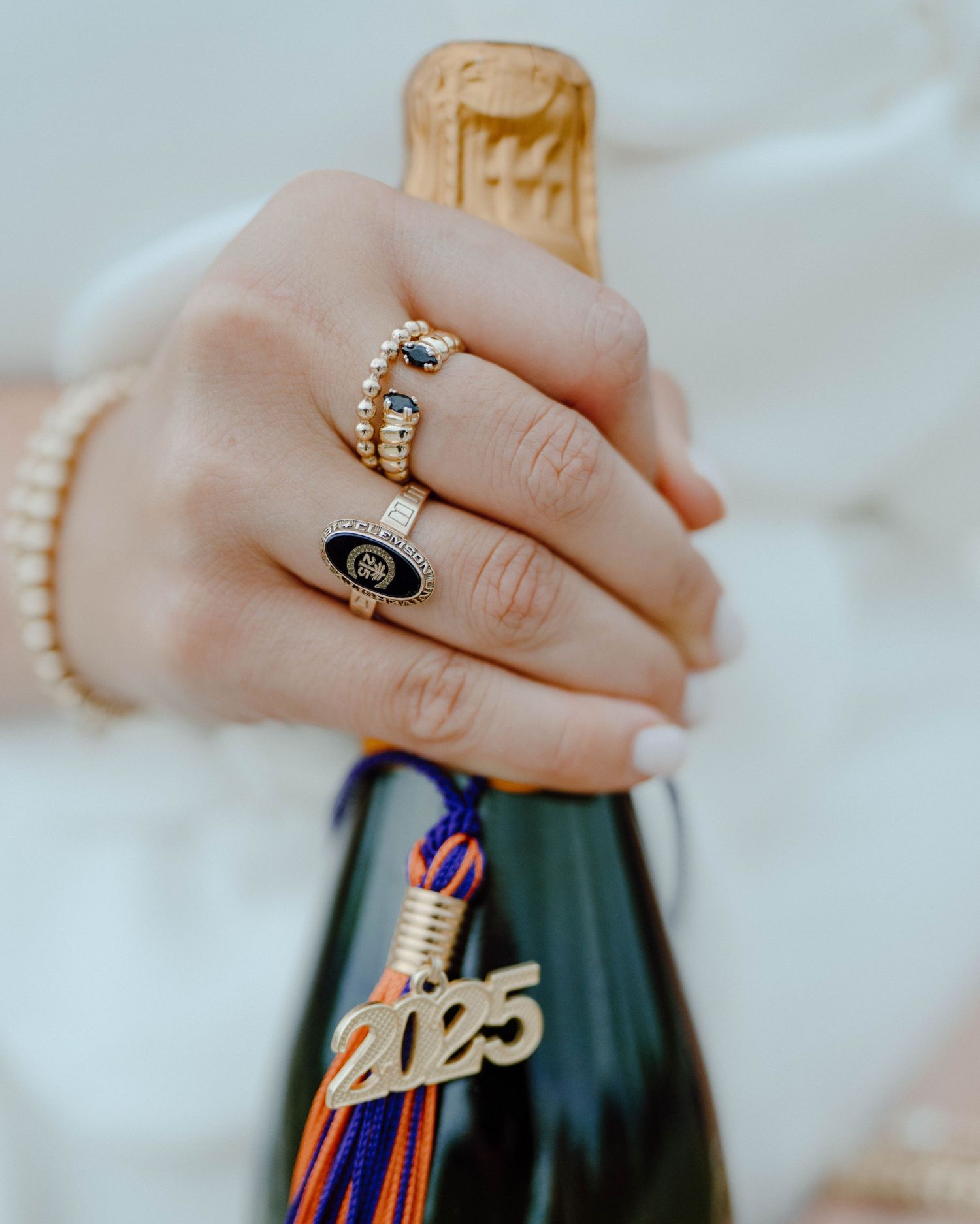 Close-up of hands wearing jewelry holding a graduation tassel and champagne bottle during celebration.