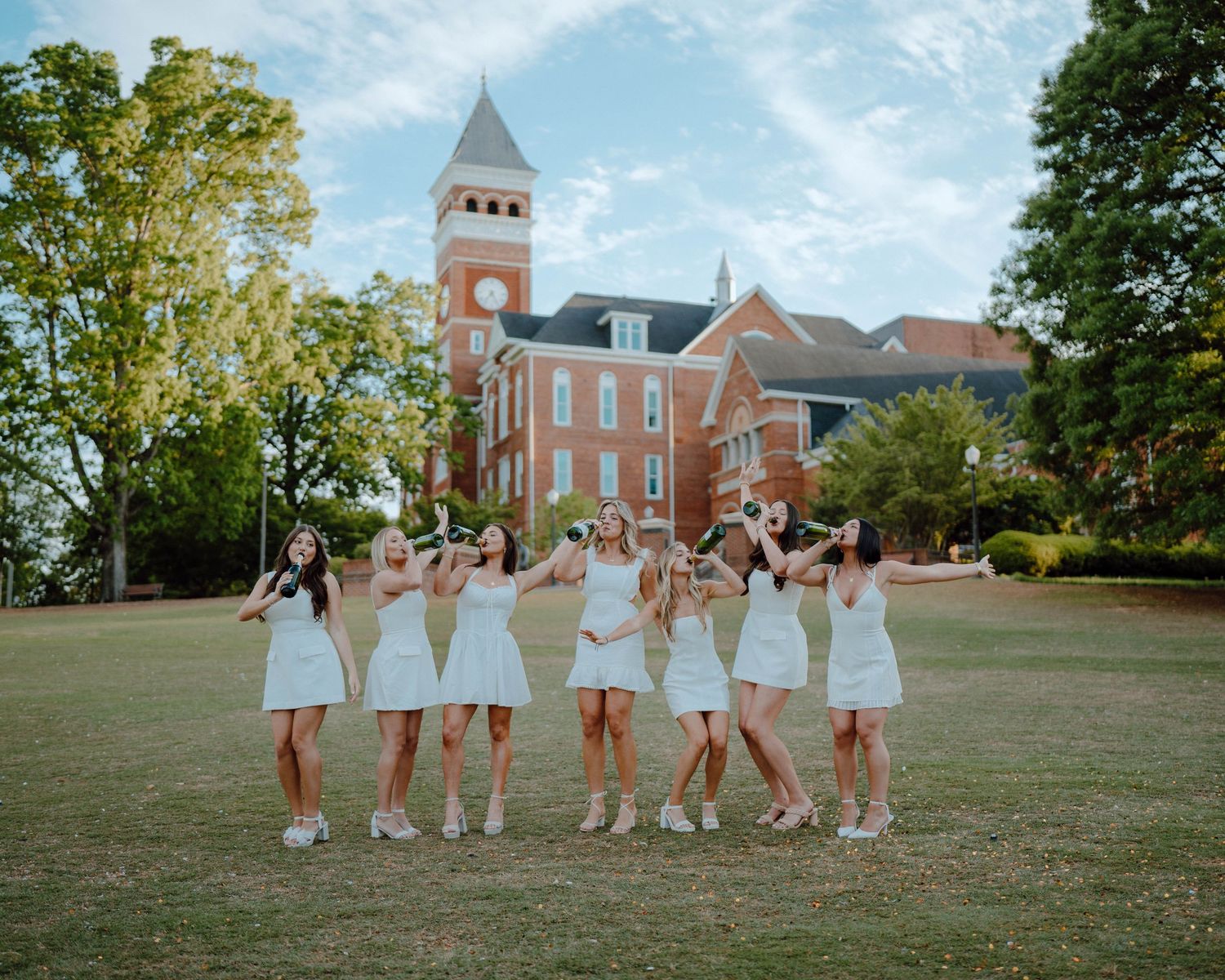 A group of friends in matching white dresses jump together on a lawn in front of a historic brick building with a clock tower.